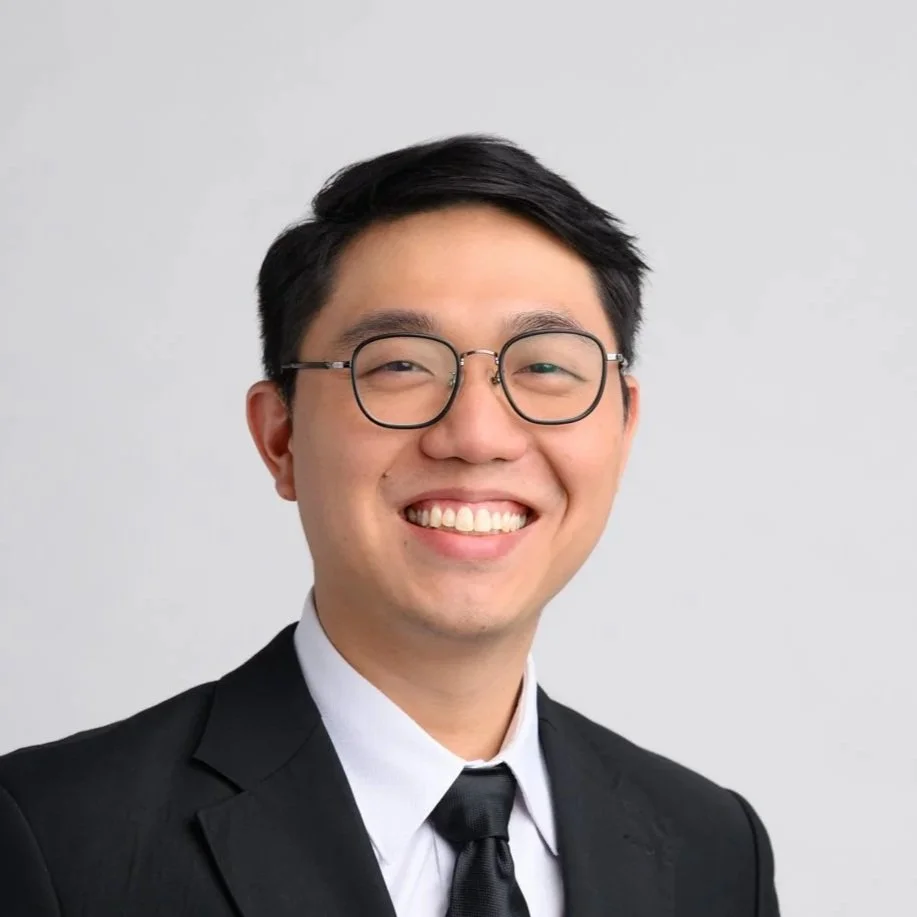Portrait of a young Asian man with black hair and glasses, smiling, wearing a black suit, white shirt, and black tie, against a plain gray background.