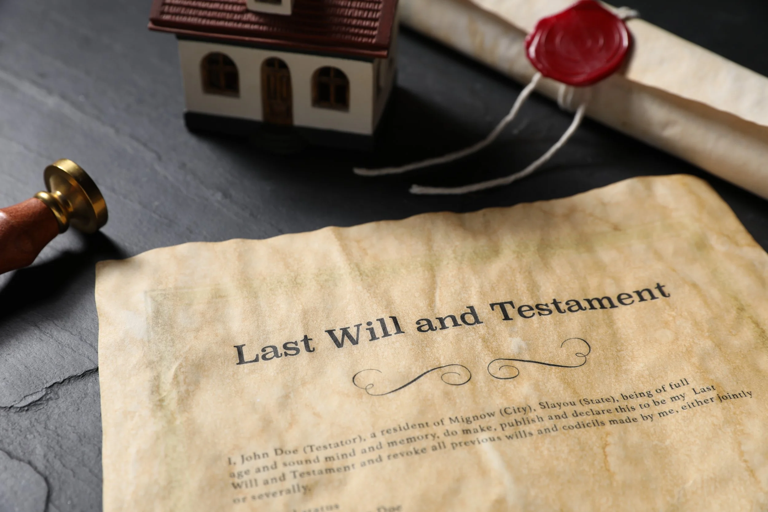 An aged and crumpled legal document titled 'Last Will and Testament' with a vintage paper appearance, placed on a dark surface. Part of a small model house, a wooden stamp handle with a gold-colored stamp top, and a rolled-up parchment with red wax seal are visible in the background.