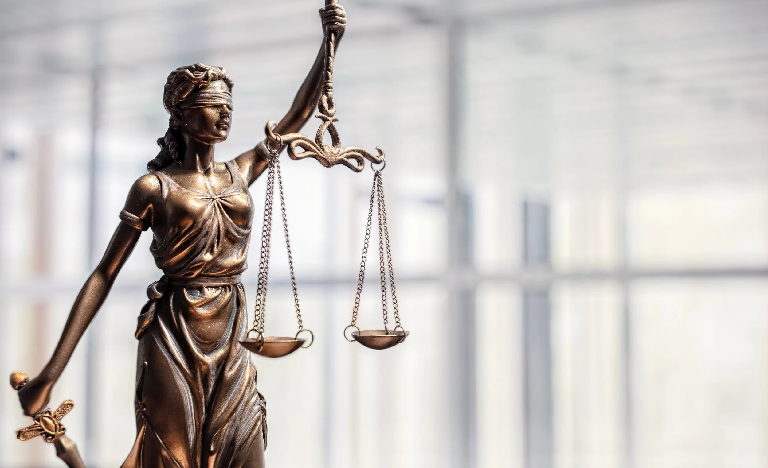 Close-up of a bronze statue of Lady Justice, holding scales of justice in her left hand and a sword in her right hand, with a blurred background of a modern court or government building.