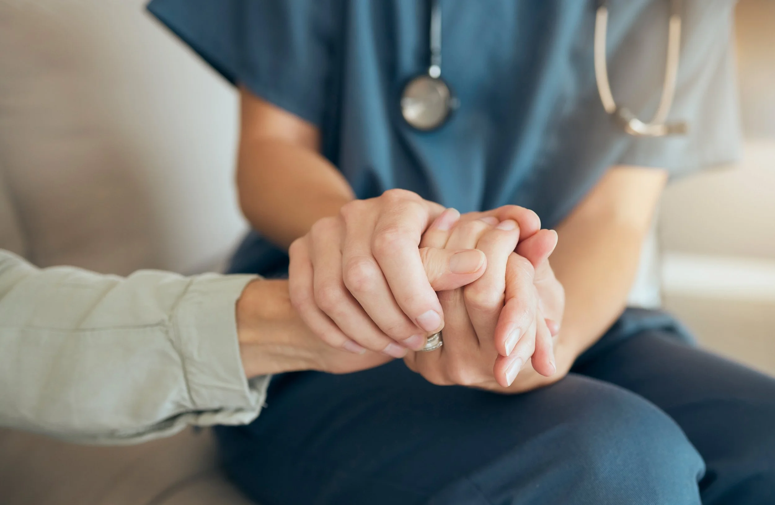 Close-up of two hands clasped in a supportive gesture, with a person wearing medical scrubs and a stethoscope around their neck in the background.