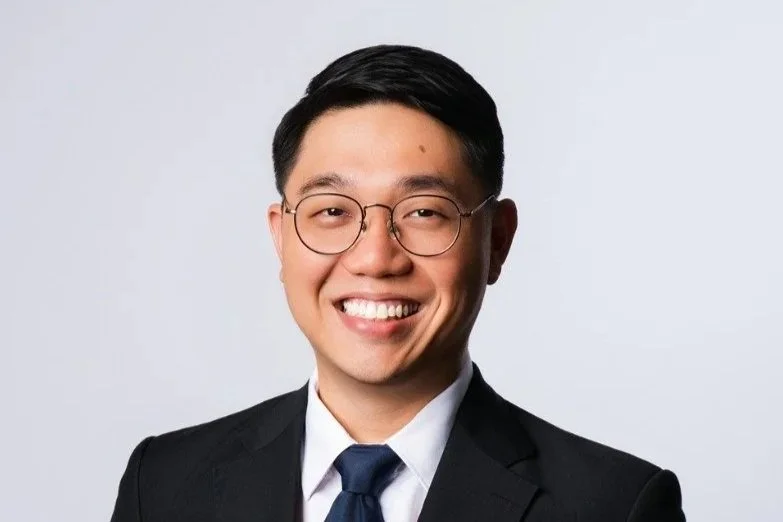 Portrait of a young man in a black suit, white shirt, and black tie, smiling with glasses, standing against a plain gray background.
