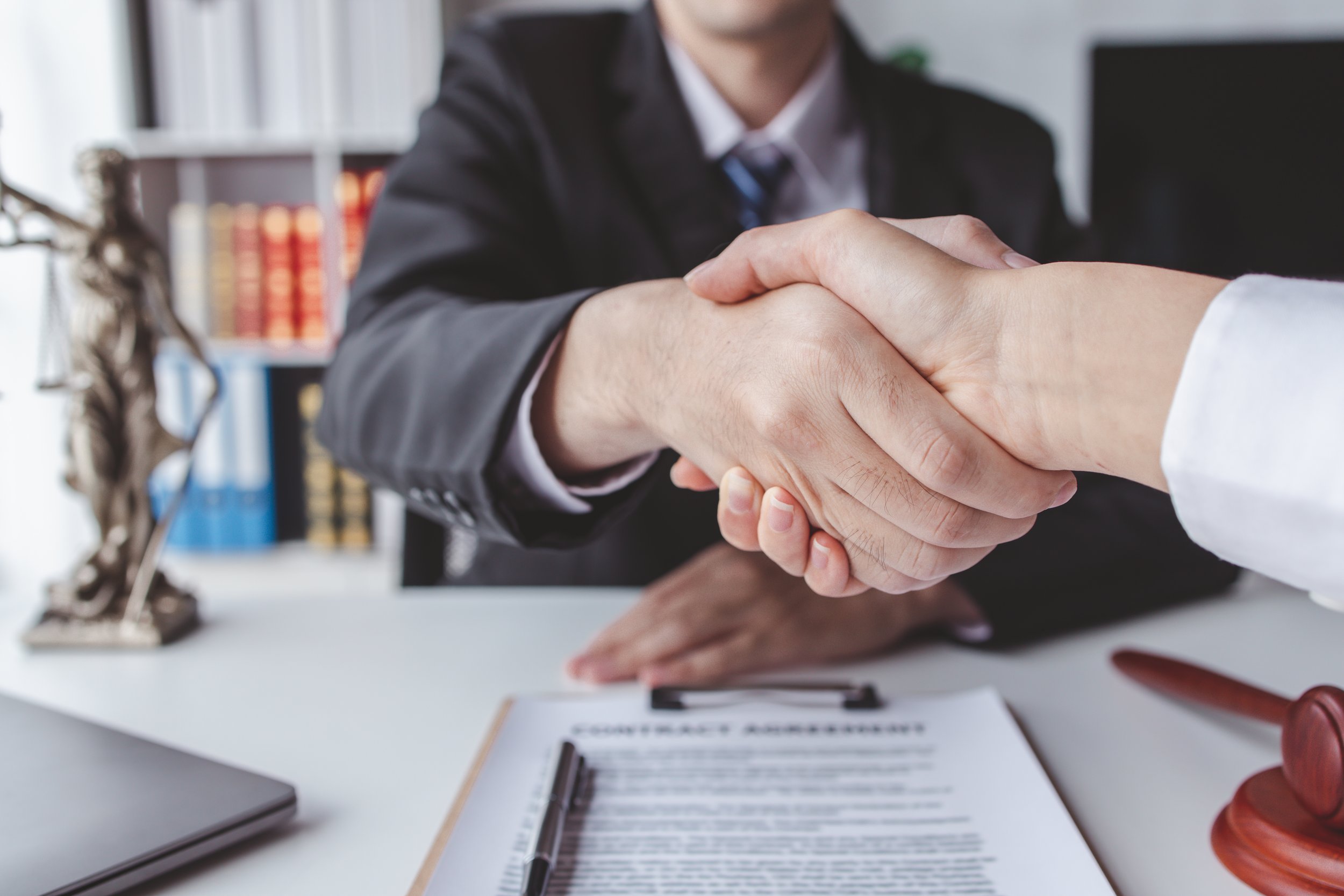 Handshake between two people, one dressed in a suit and the other in a white shirt, in an office setting with legal documents, a gavel, and a scale of justice on the desk.