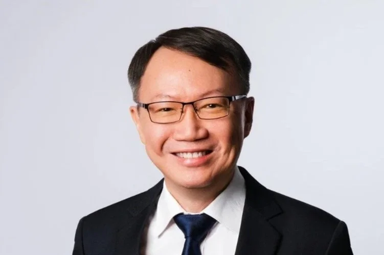 Professional headshot of a man with glasses, wearing a suit and tie, smiling against a white background.