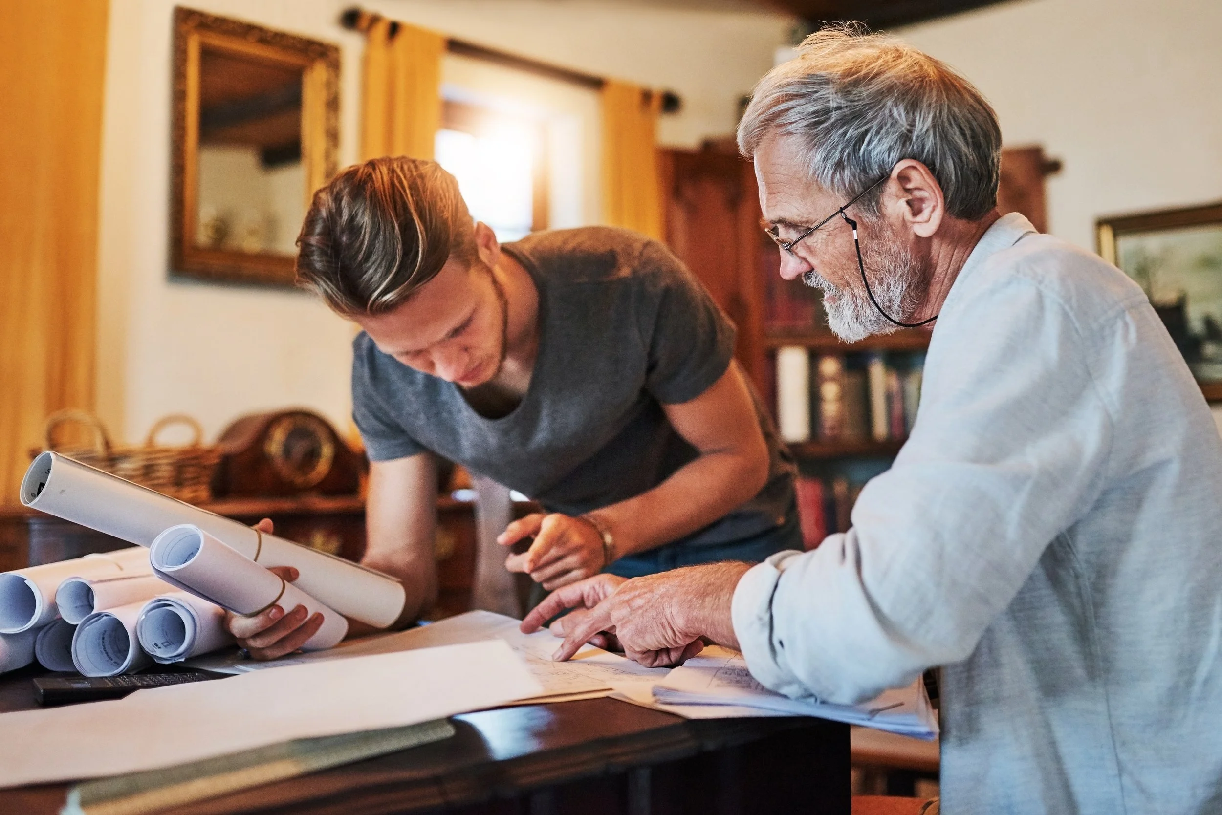 Two men are examining blueprints and rolled-up architectural plans on a work table in a room with wooden furniture and yellow curtains.