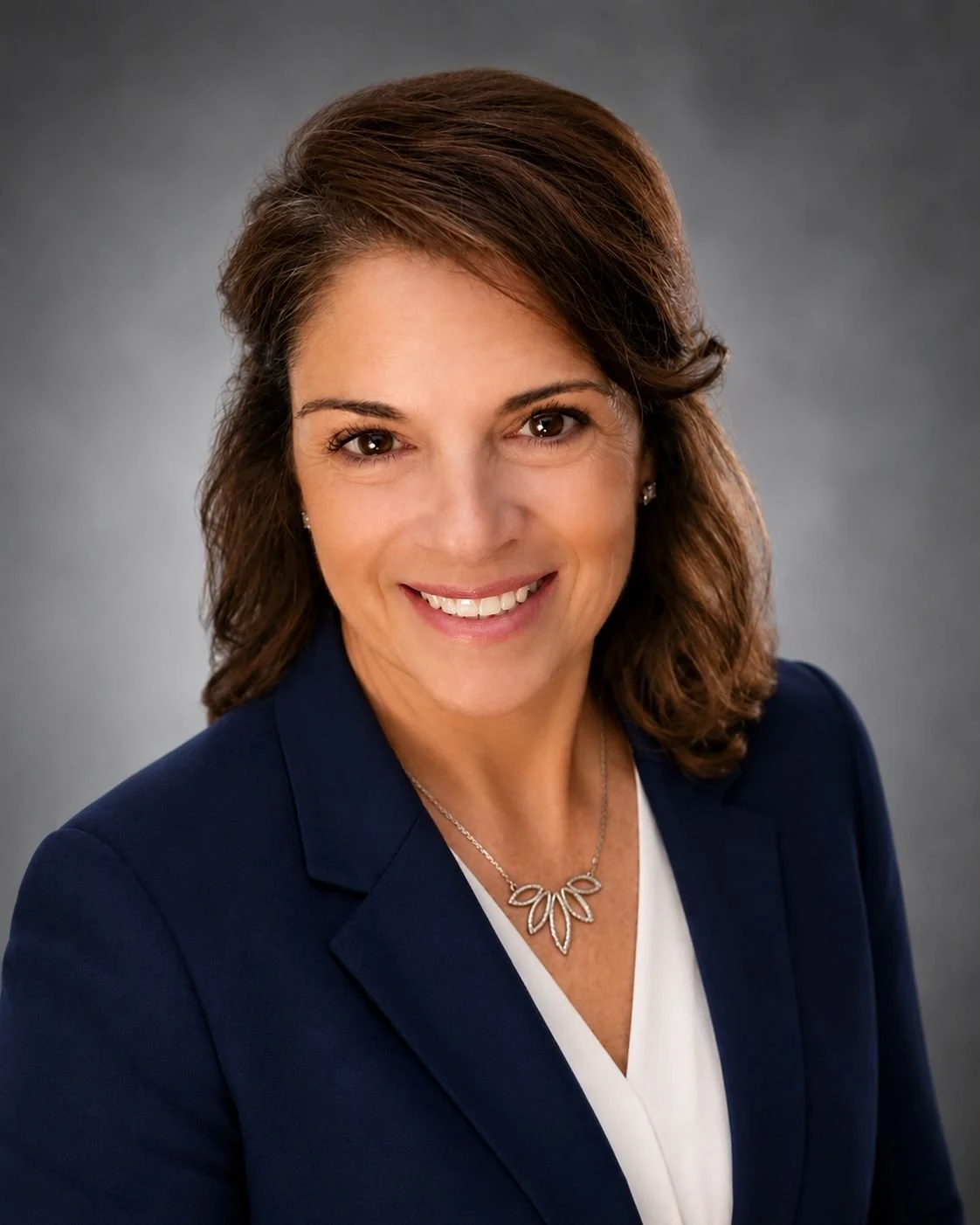 Professional woman with brown hair and brown eyes smiling, wearing a navy blazer, white top, and a silver leaf-shaped necklace, against a gray background.