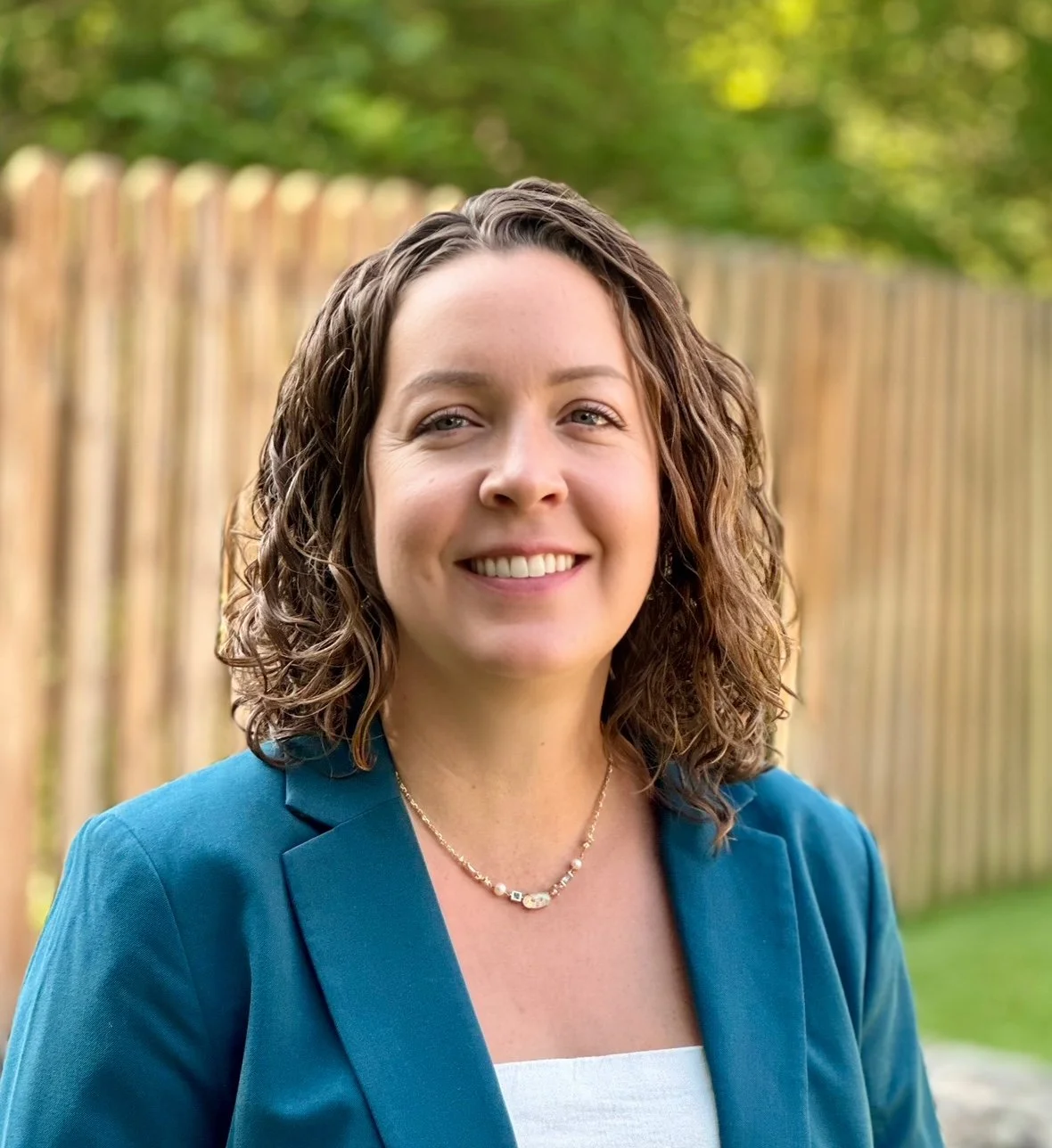 A smiling woman with shoulder-length curly brown hair, wearing a blue blazer and a pearl necklace, standing outdoors with a wooden fence and green trees in the background.