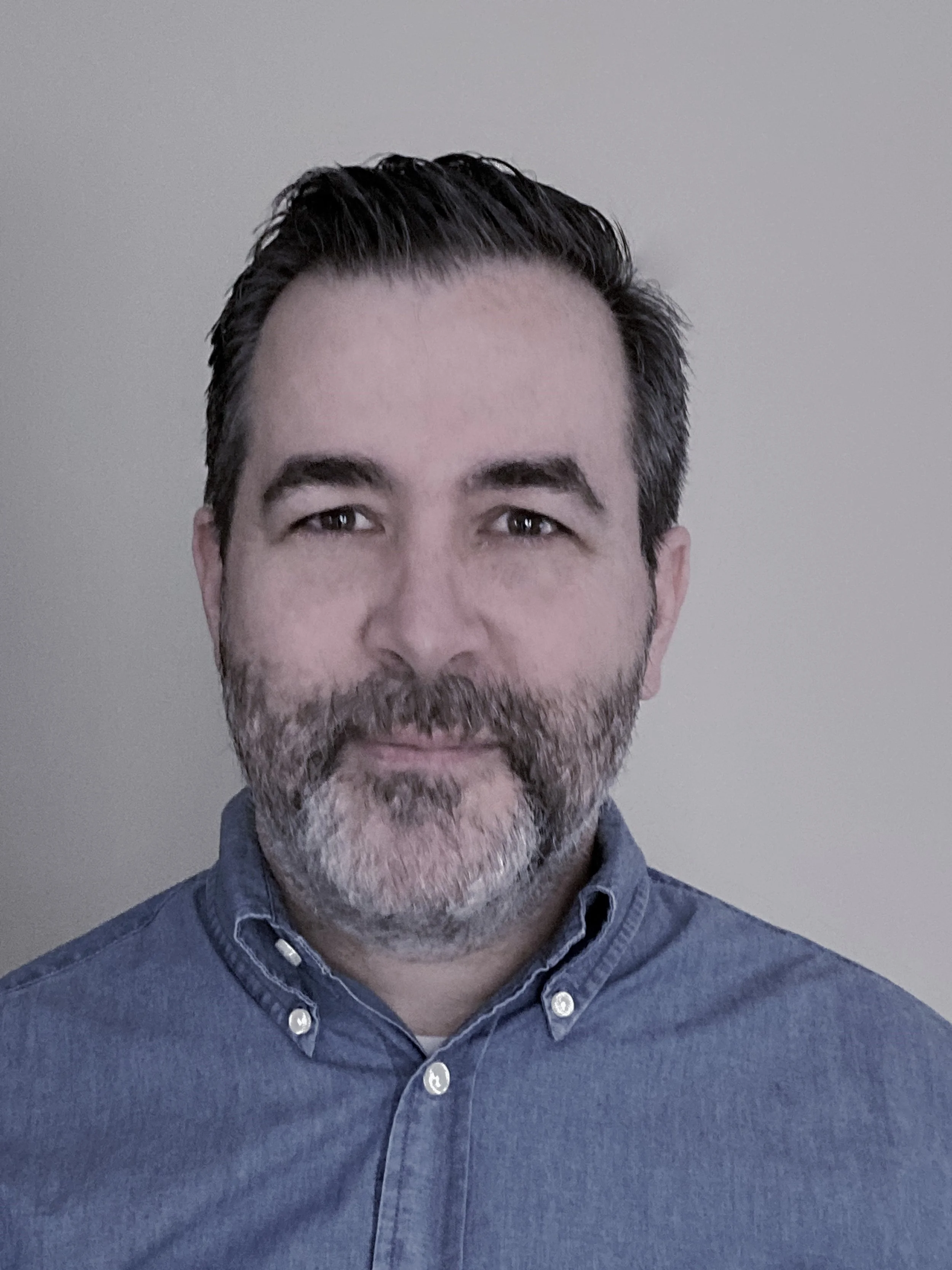 Close-up of a man with dark hair and a beard, wearing a jean button-up shirt, against a plain light-colored wall.