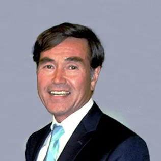 A man with dark hair, wearing a dark suit, white shirt, and a light blue tie, smiling and posing against a plain background.