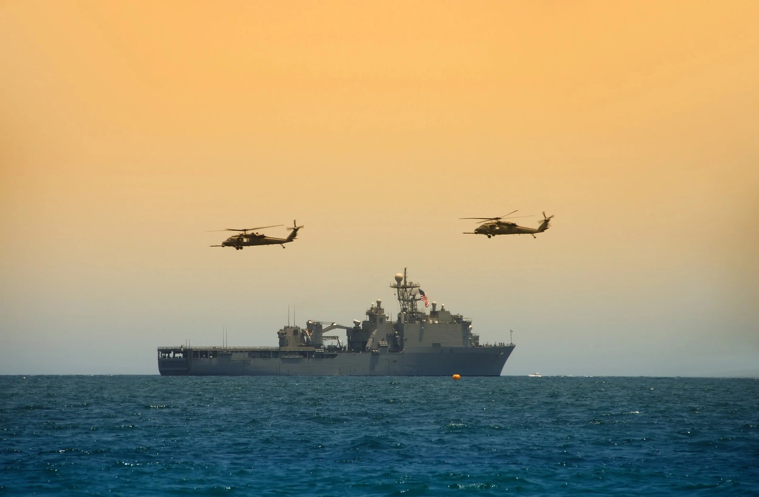 Two military helicopters flying in formation above a navy ship at sea during sunset.
