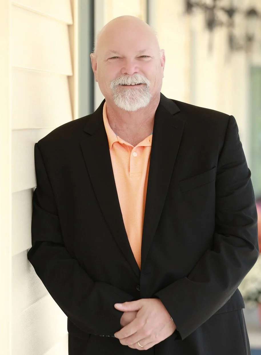 A man in a black suit jacket and peach-colored polo shirt standing outdoors near a building with cream-colored siding, smiling at the camera.