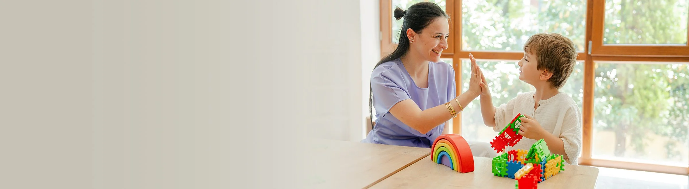 A woman and a young boy at a table playing with colorful plastic building blocks, giving each other a high-five, with a rainbow-shaped model on the table, and a large window in the background showing greenery.