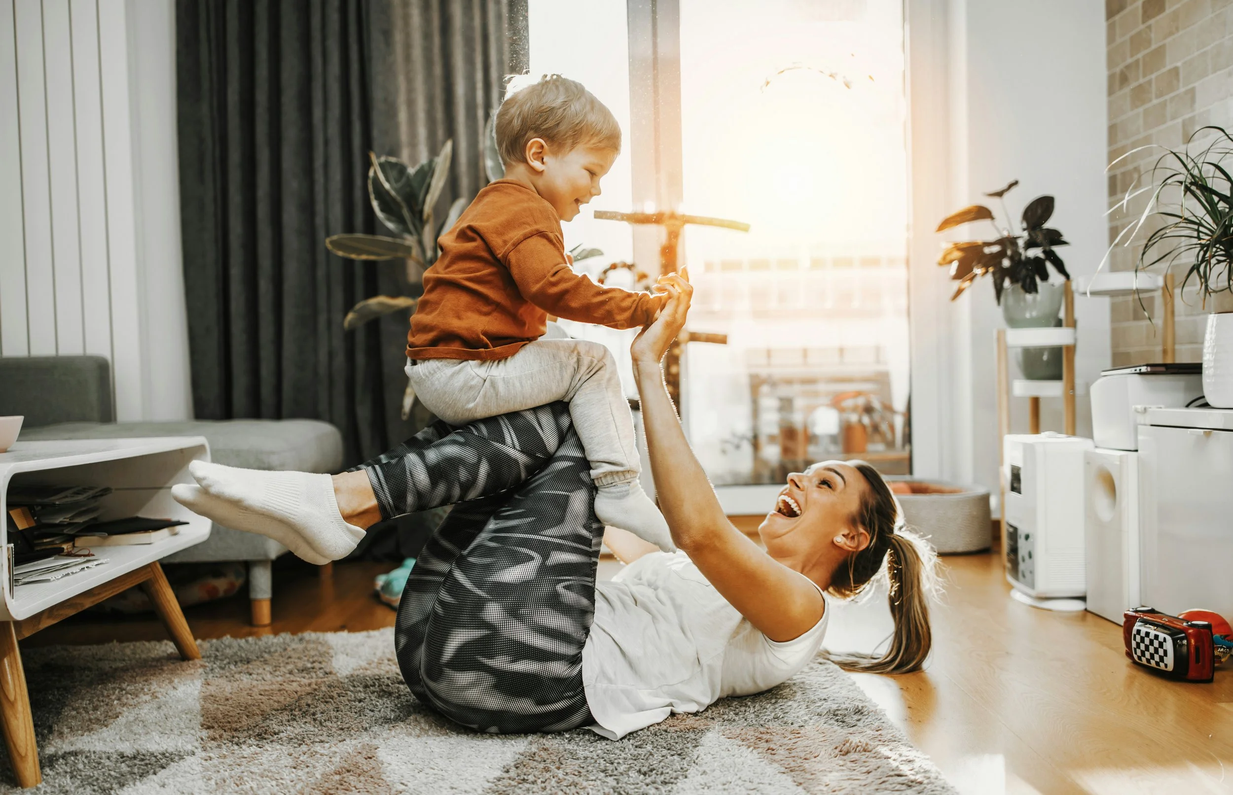 A woman and a young boy playing on a living room carpet, with the woman lying on her back and lifting the boy into the air as he smiles and reaches out.
