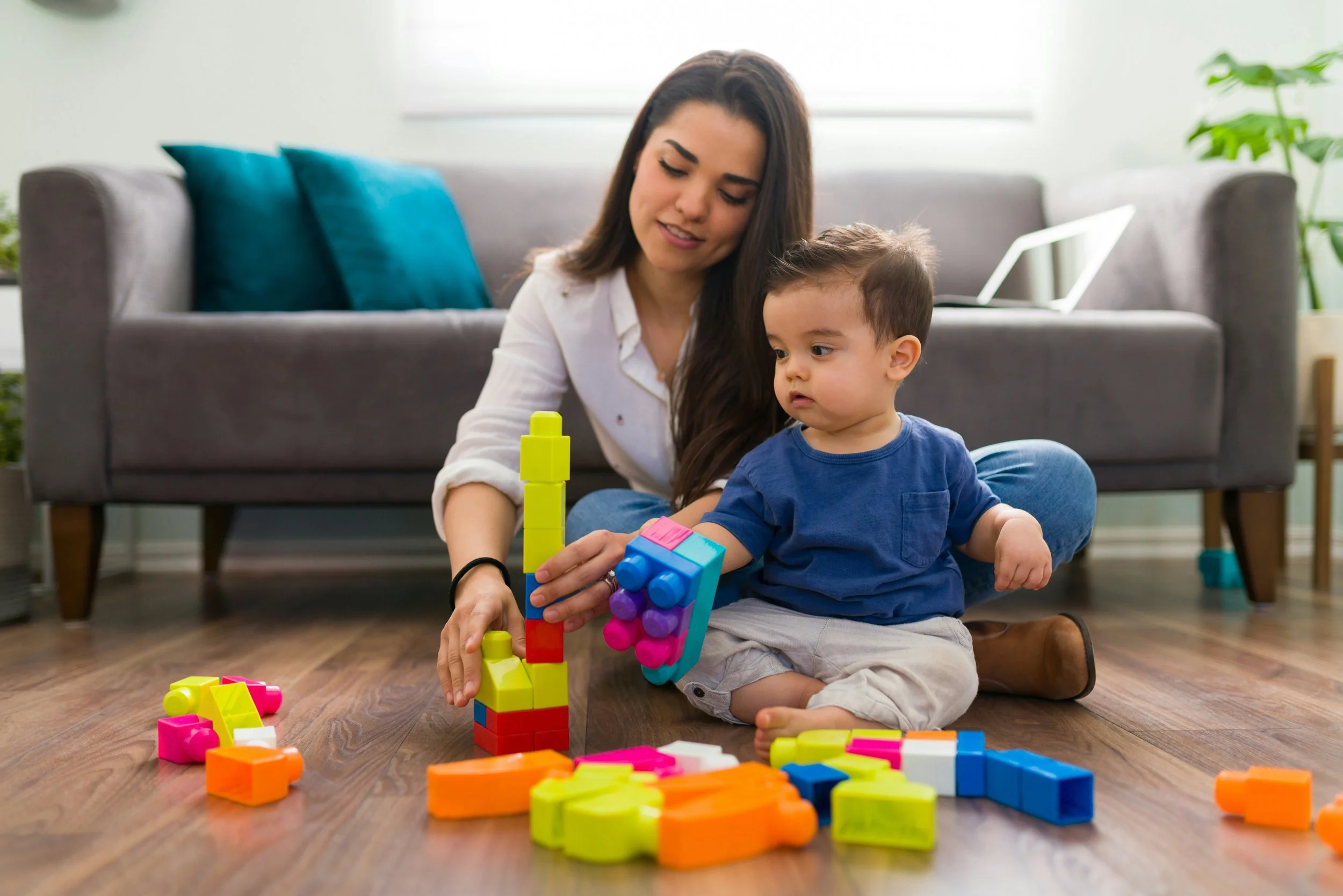 A woman and a young boy sitting on the floor playing with colorful building blocks indoors, with a sofa, pillows, and a laptop in the background.