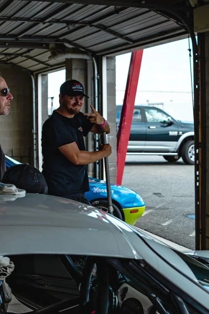 A man in a black t-shirt and cap giving a peace sign next to a blue race car in a garage. Another person is partially visible on the left, and a dark vehicle is outside the garage.
