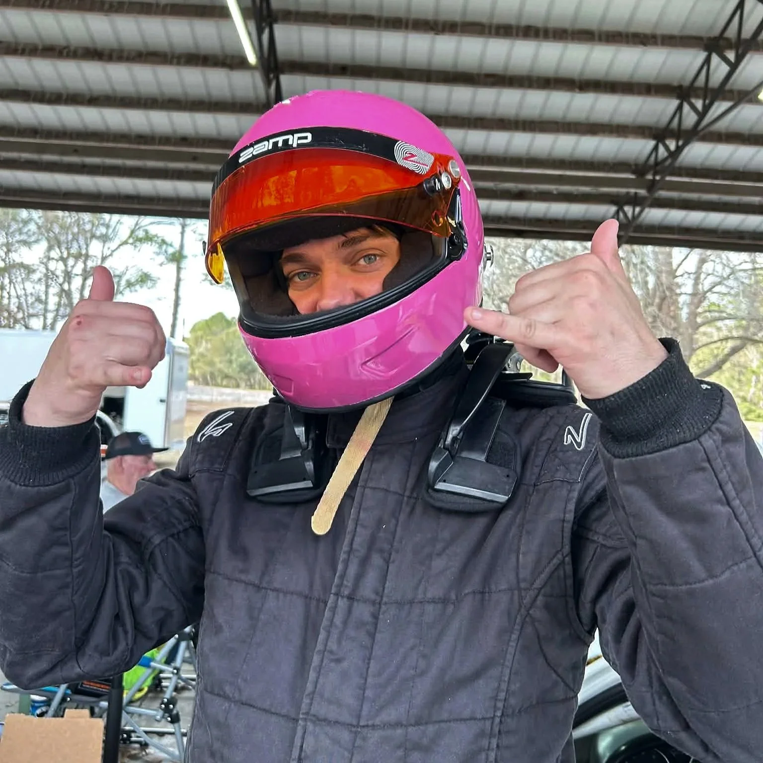 A man in a racing suit and a pink helmet giving a thumbs-up while talking on a phone at a racing track.