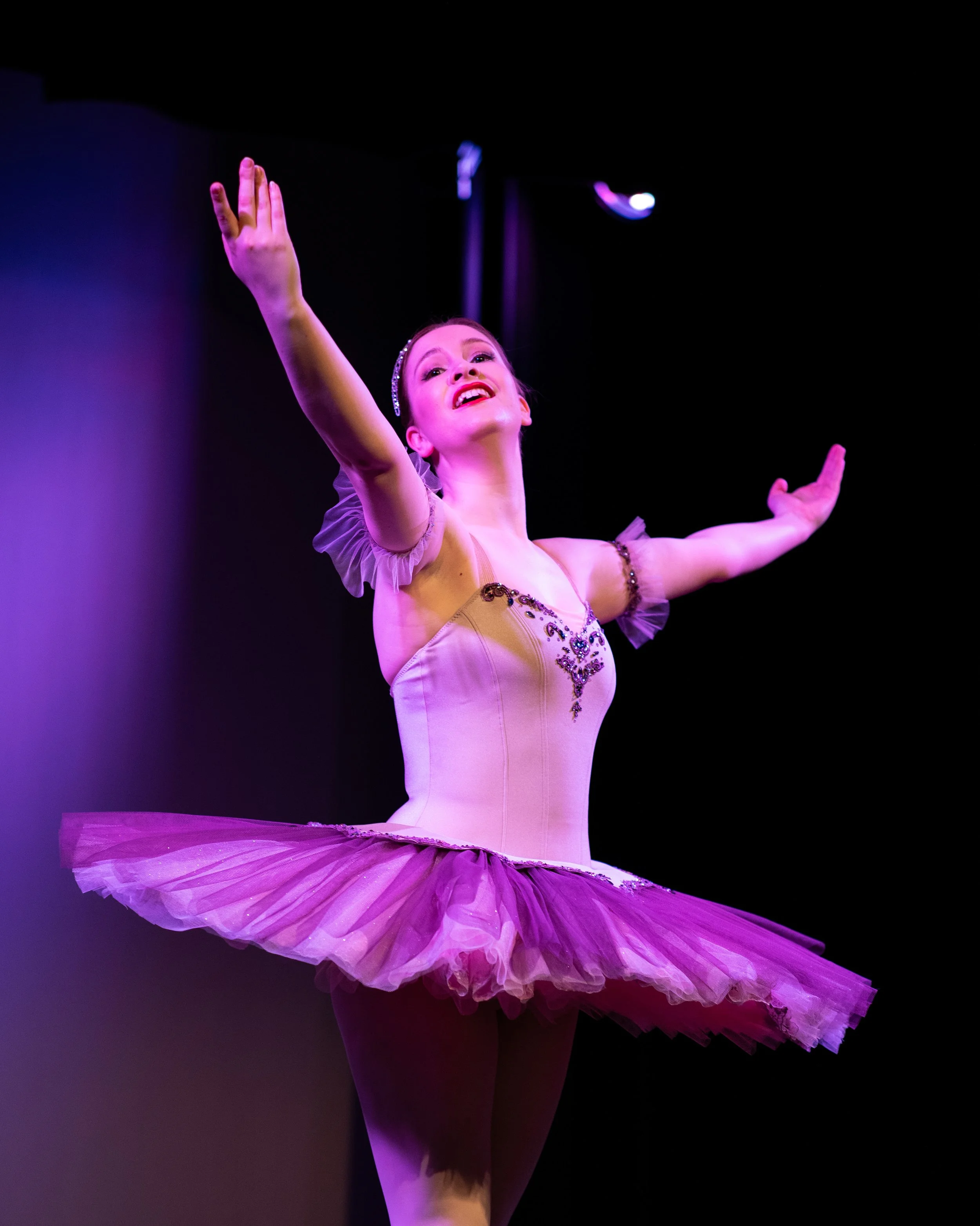 Adult Ballet Centre dancer Rhi as a ballerina in pink tutu dancing on stage with arms raised, purple stage lighting for the performance season