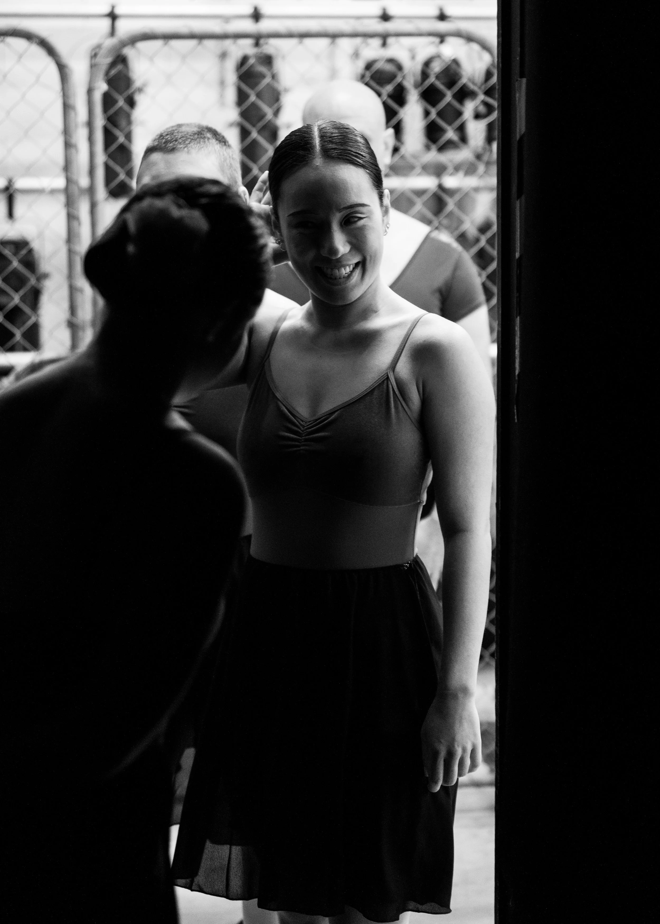 Adult Ballet Centre dancer Rebecca with dark hair tied back, smiling, standing back stage with a chain-link fence backstage  and other people in the background, in black and white.