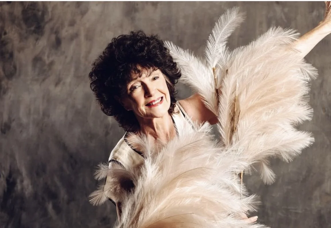 Anne Marie Dowling Adult Ballet Centre Patron photographed with curly dark hair holding large beige feathers, smiling, neutral background.