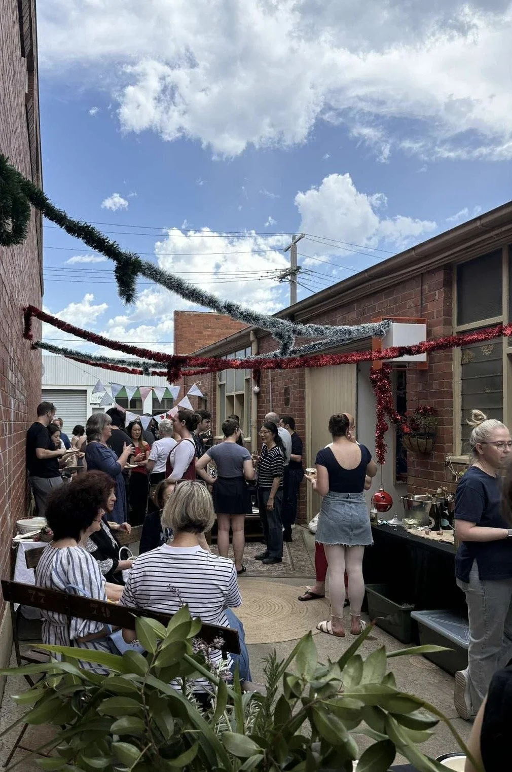 People gather in a small outdoor laneway for the Adult Ballet Centre Christmas Party  with brick walls, holiday decorations, string lights, and picnic tables, enjoying a festive social event on a sunny day.