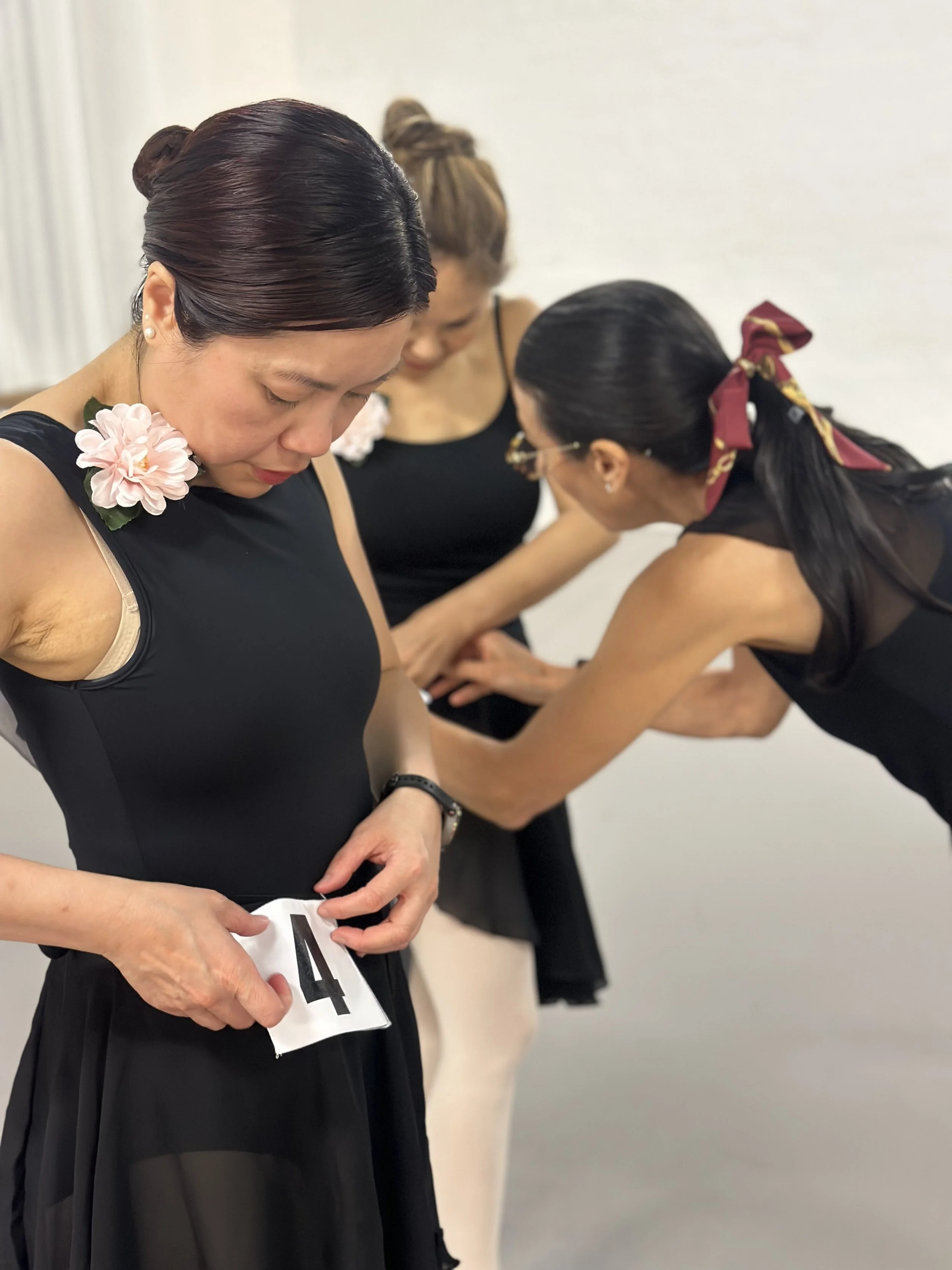 Three women in black dresses are preparing for a competition, with one woman holding a number tag and two women adjusting her outfit.