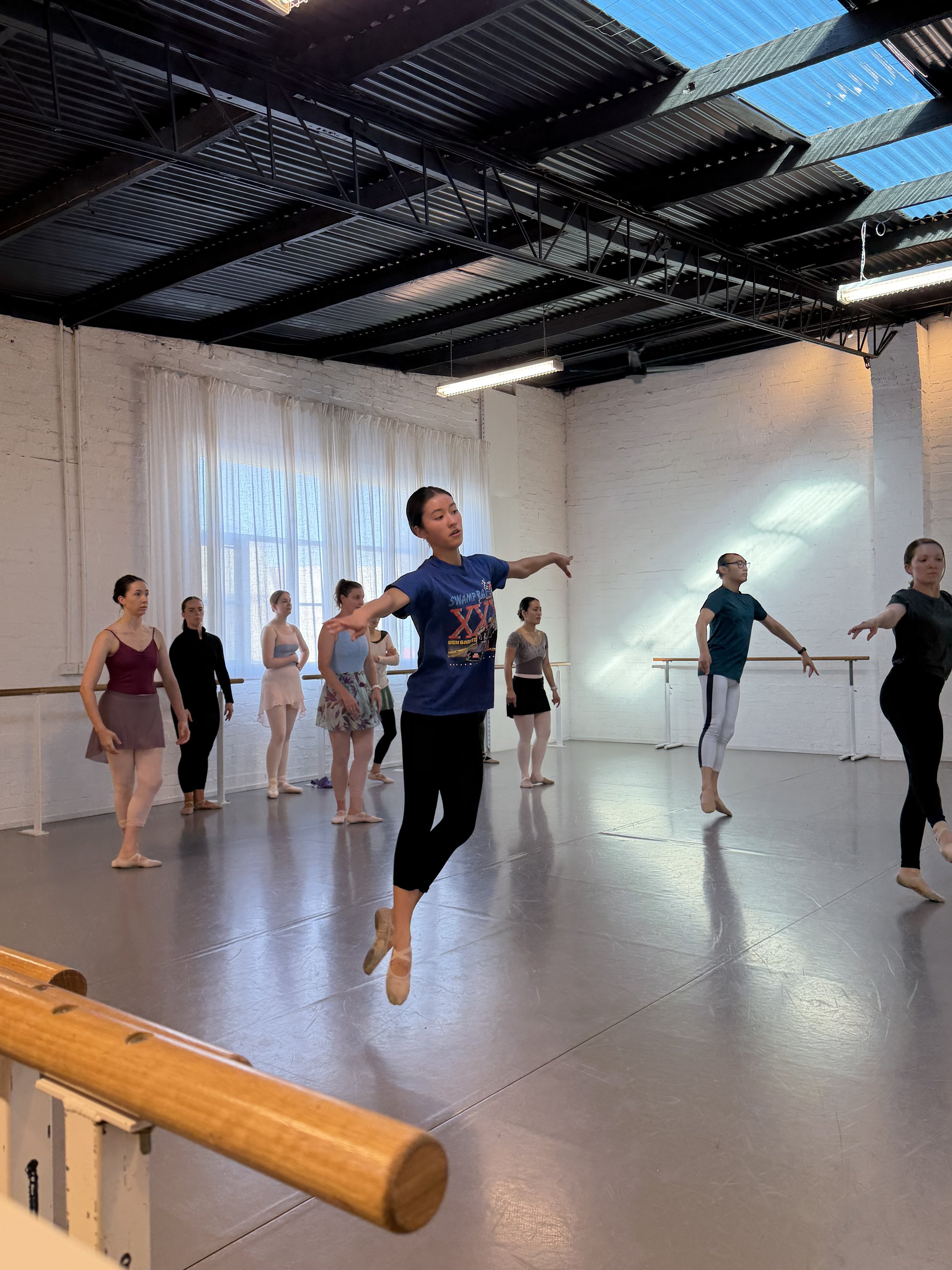 Adult Ballet Centre dancers practicing in a dance studio with white brick walls, large windows with curtains, wooden barre, and black ceiling with exposed metal beams.