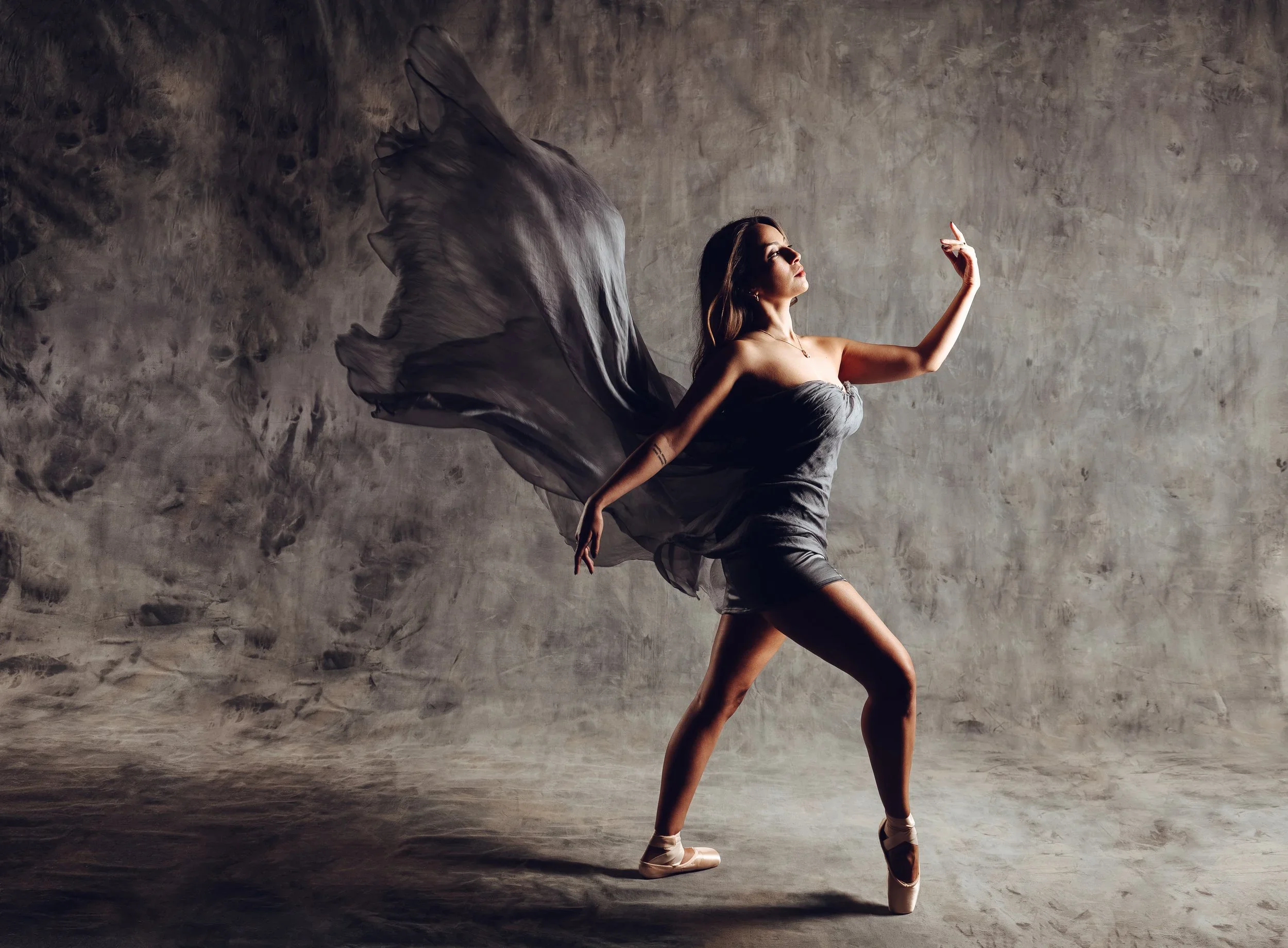A woman dancing ballet on a studio floor, wearing a gray dress and pointe shoes, with a flowing gray fabric extending from her back, against a textured gray wall.