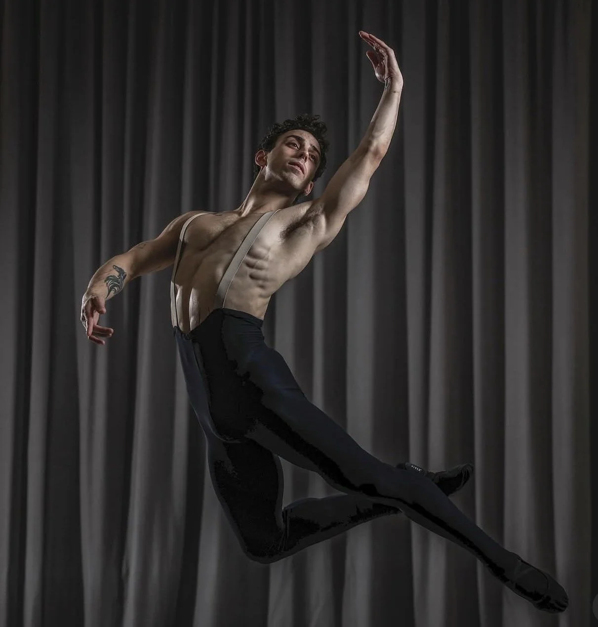 A male ballet dancer performing an arabesque on stage, dressed in black tights and suspenders, against a dark curtain backdrop.