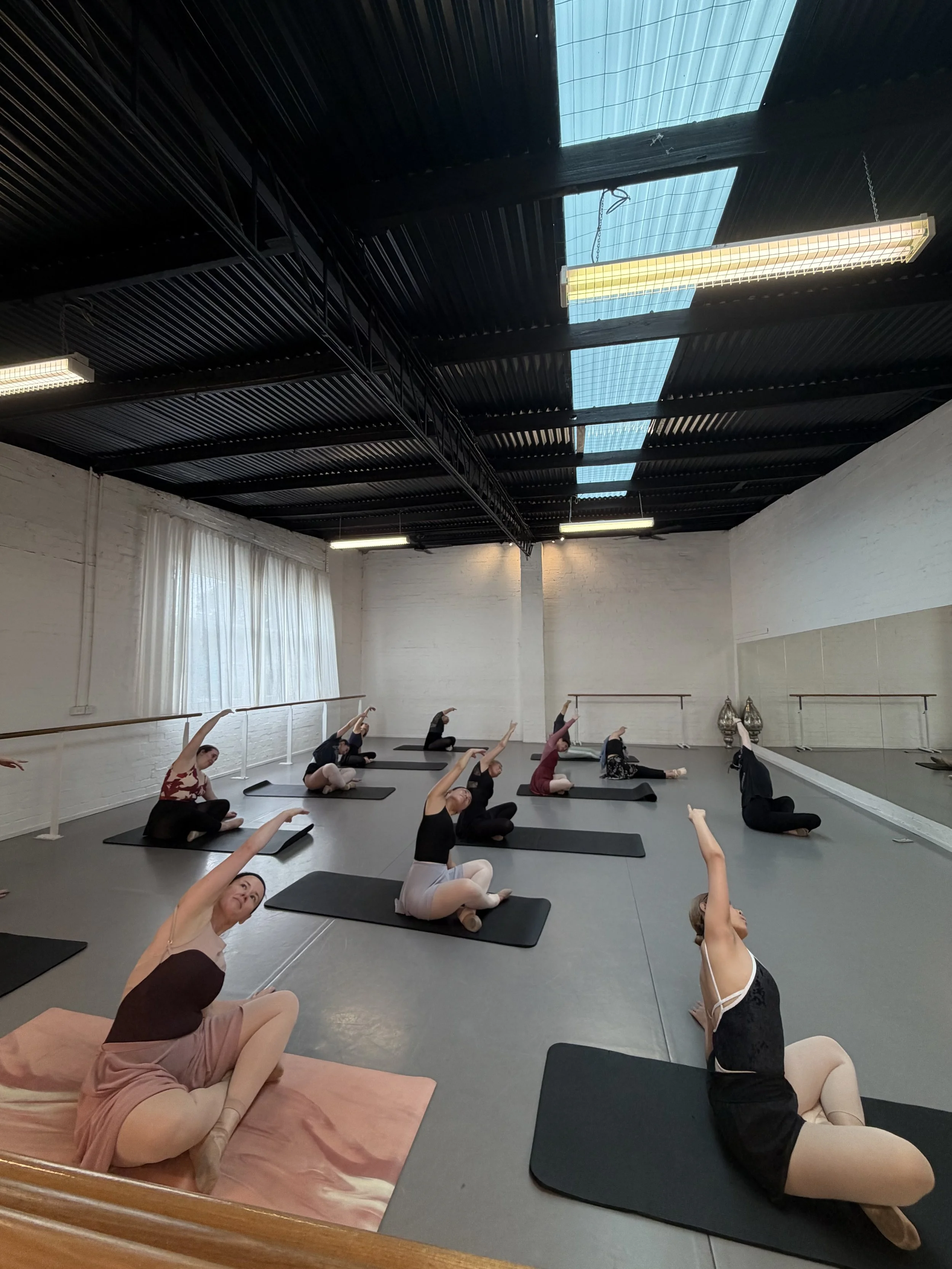 Group of people practicing yoga in a studio with mirrored wall, white curtains, and skylights in the ceiling.