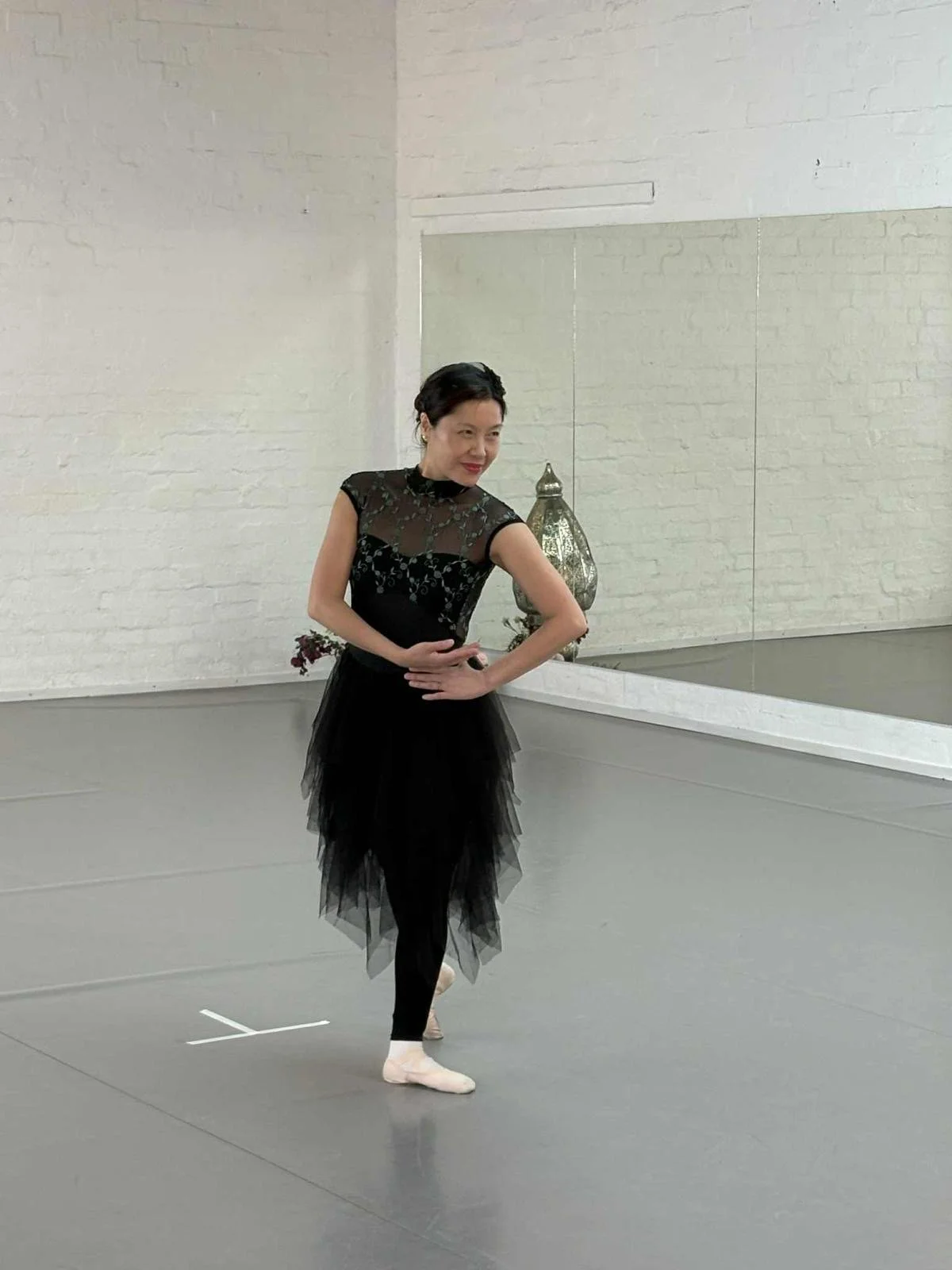 Adult Ballet Centre dancer in a black dress with a tulle skirt and lace top practicing solo ballet private lesson in the Adult Ballet Centre dance studio with white brick walls and a mirror.
