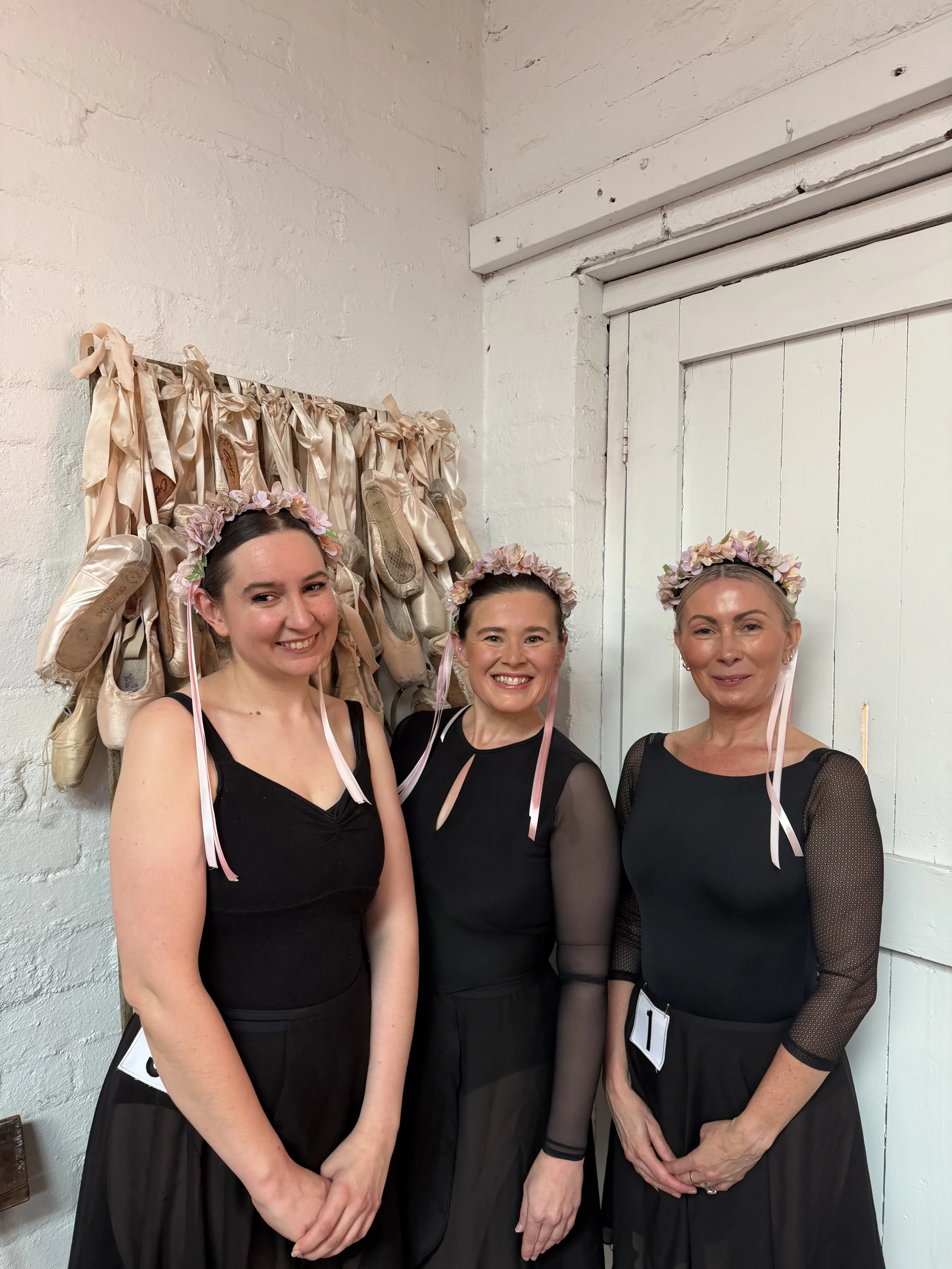 Three women wearing black dresses and floral headbands smiling and standing together in front of a white wall with ballet shoes hanging on a rack.