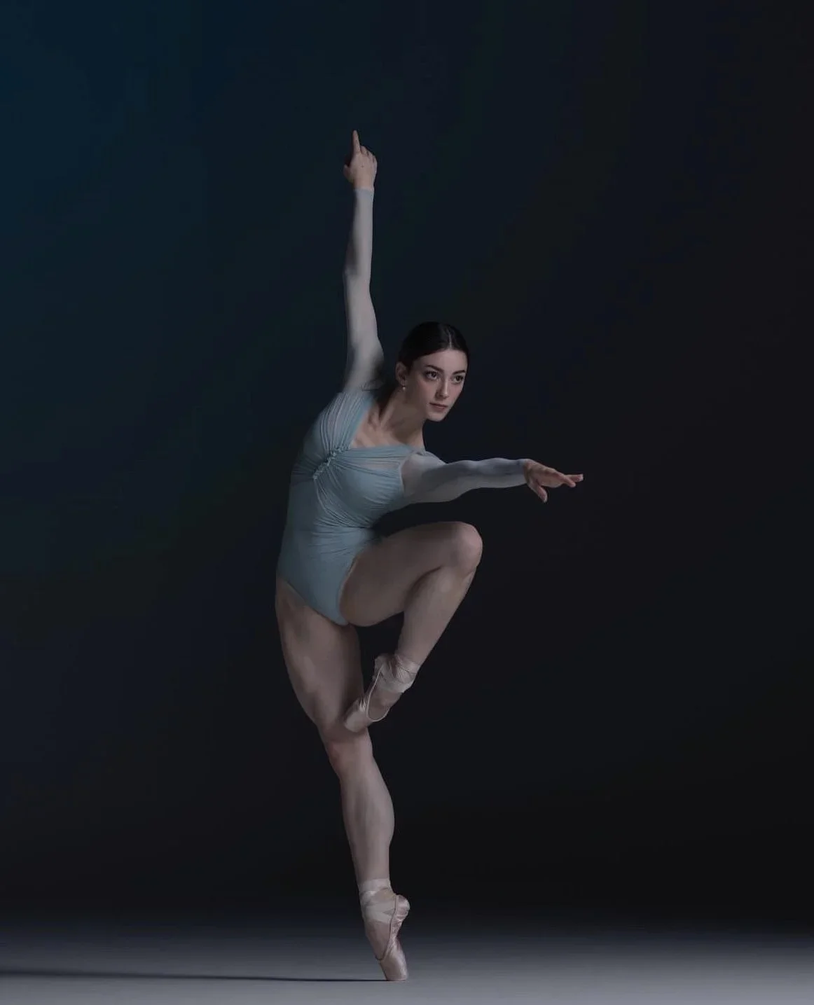 A female ballet dancer in a gray leotard performing a graceful pose on pointe shoes against a dark background.