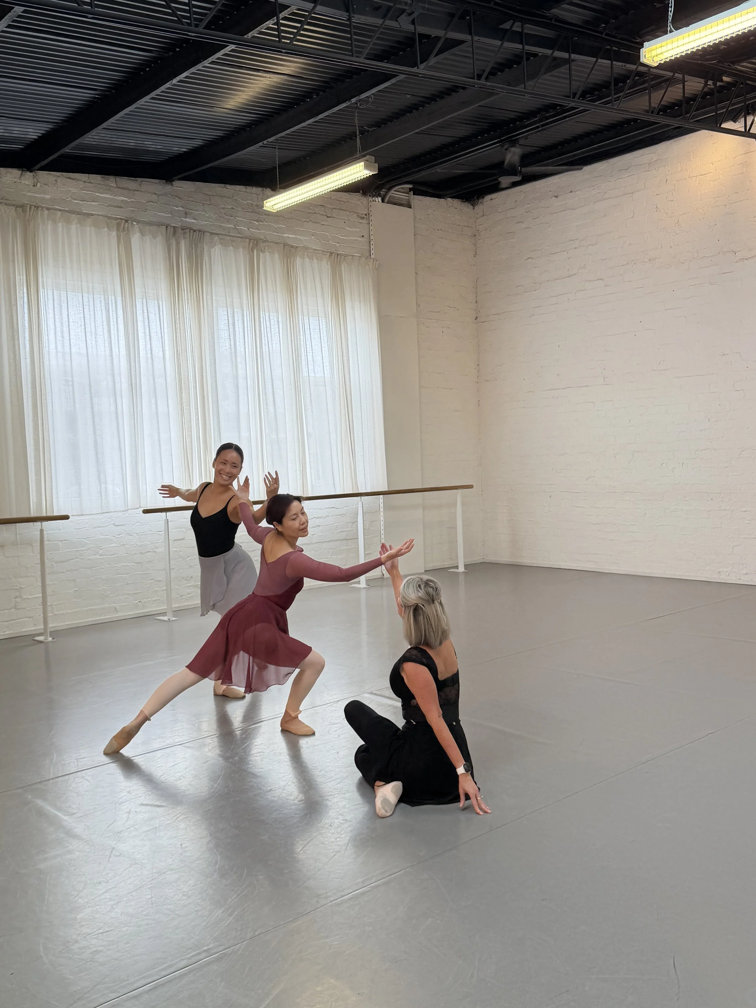 Group of ballet dancers practicing in a dance studio with white brick walls and large windows with sheer curtains, some dancers are on the floor, one is standing at the barre, and a couple of dancers are in a pose.