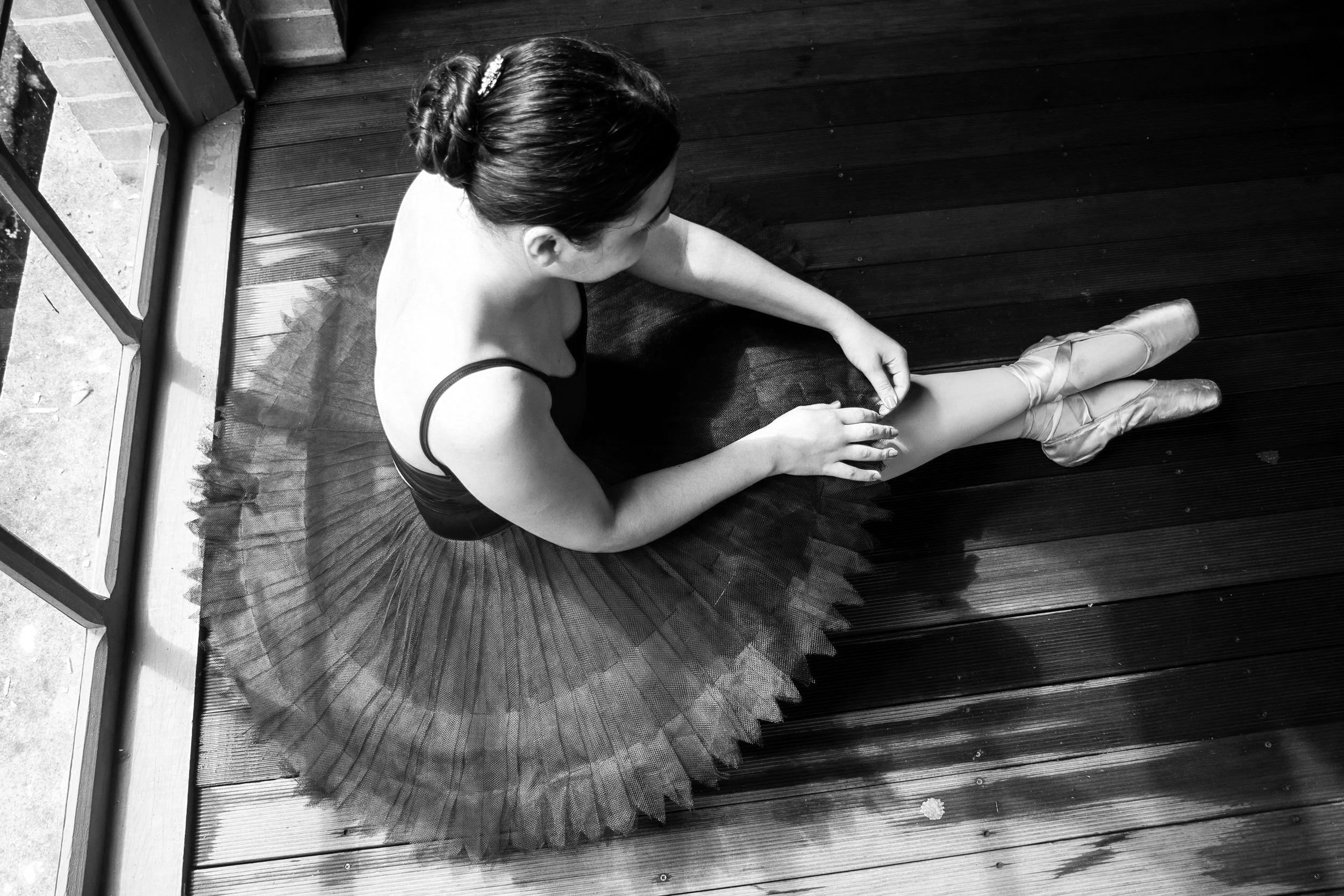 A ballerina in pointe shoes sitting on a wooden floor, wearing a tutu, with her hair styled in a bun and a hair accessory, sitting near a large window with blinds.