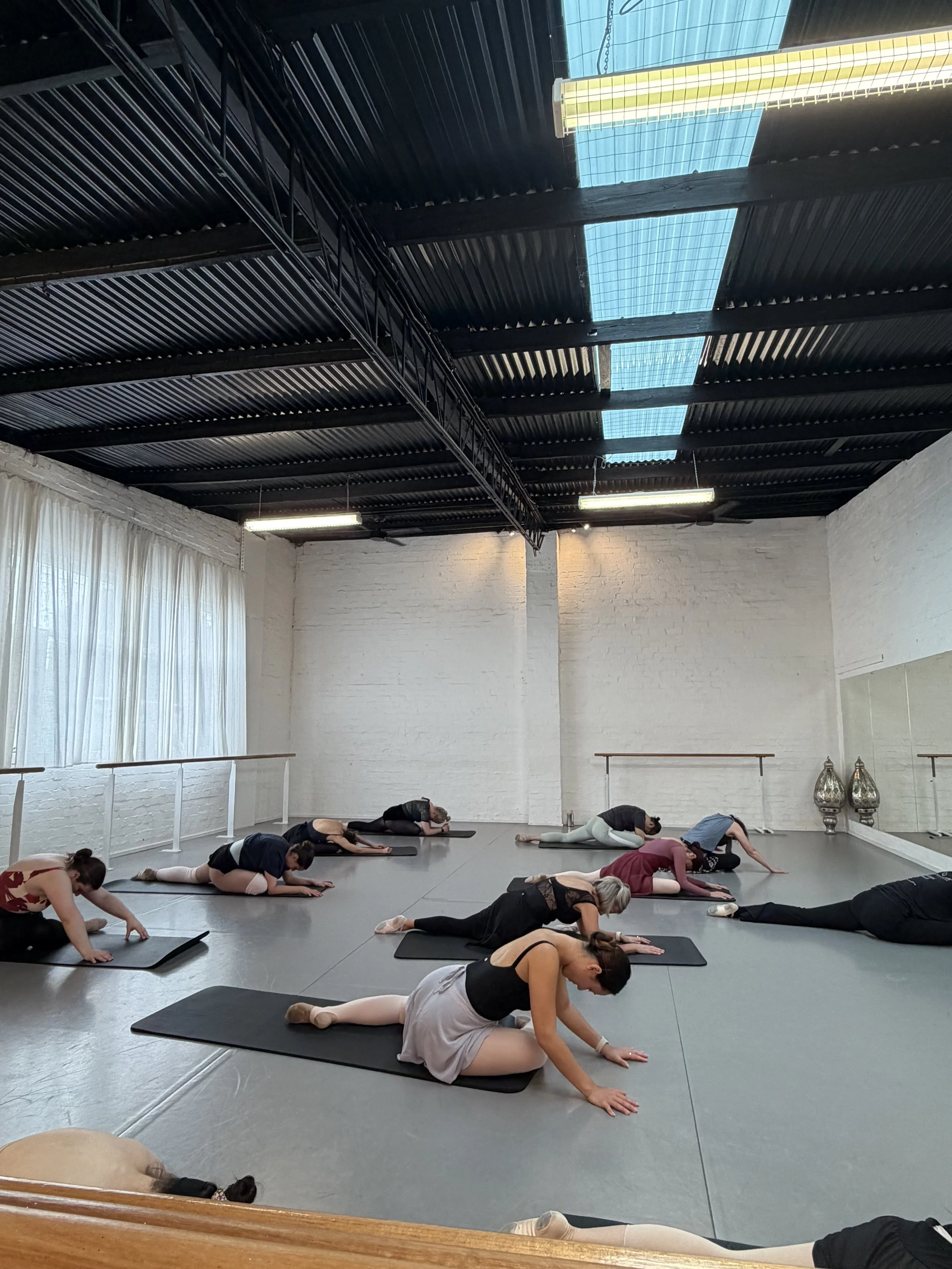 Group of people practicing yoga in a dance studio with large mirrors and a ballet barre, lying on mats in a prone position with hands extended forward.