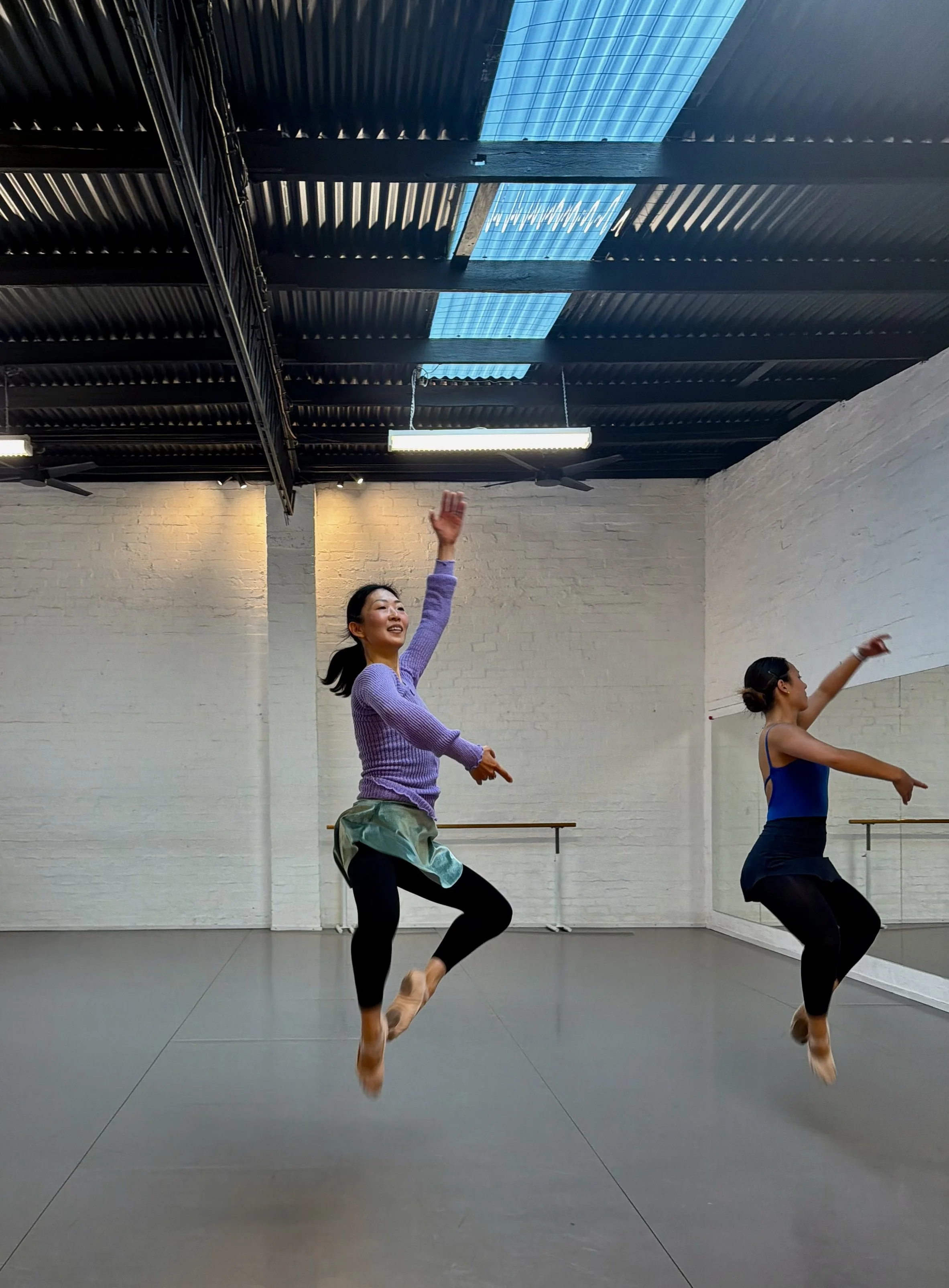Two Adult Ballet Centre dancers in a dance studio with white brick walls, mirror, and black ceiling with skylights, wearing dance attire, mid-spin with movement and grace.