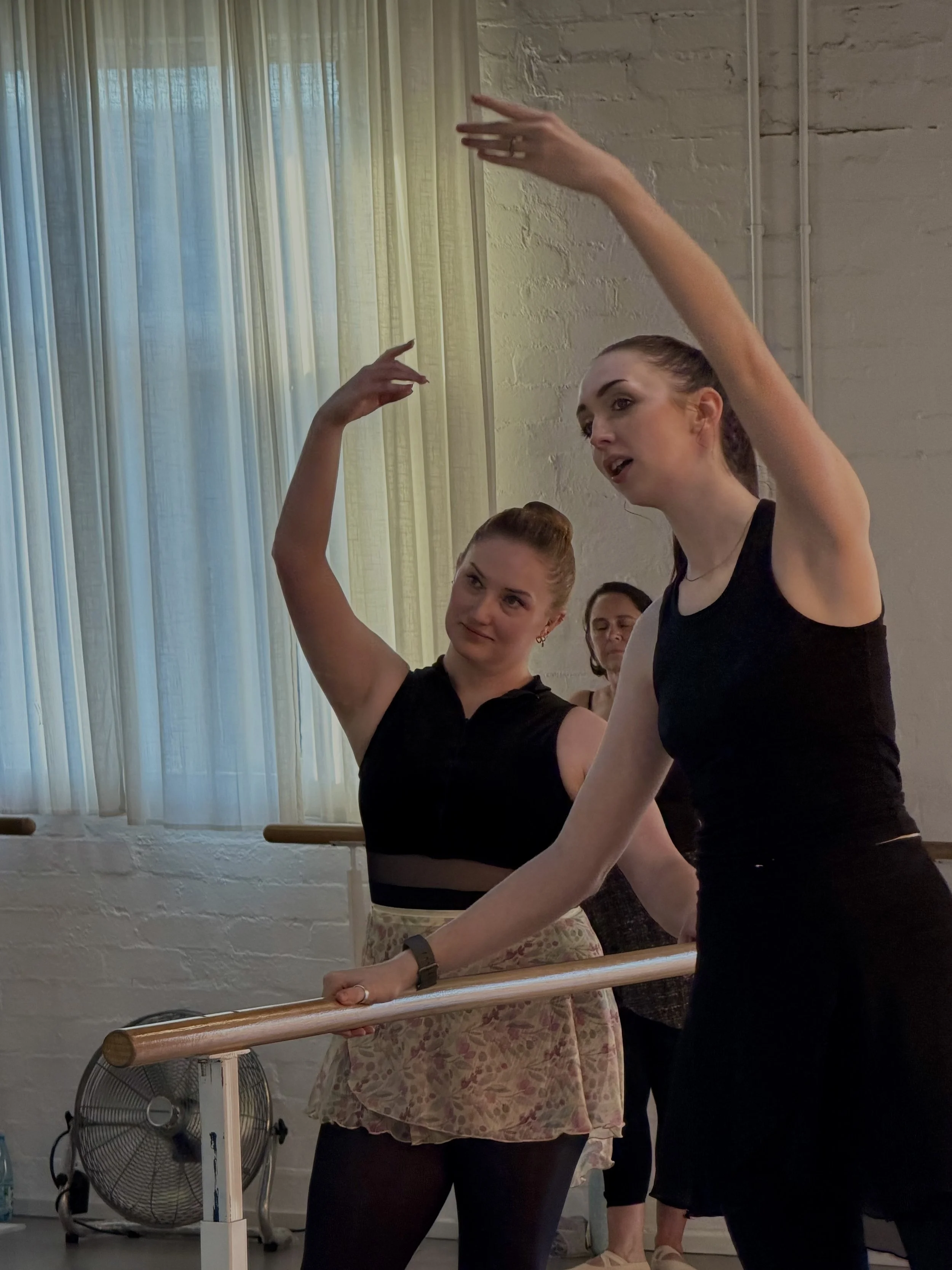 Two women practicing ballet at a dance studio, holding onto the barre, with a third woman visible in the background, and a fan at the bottom left.