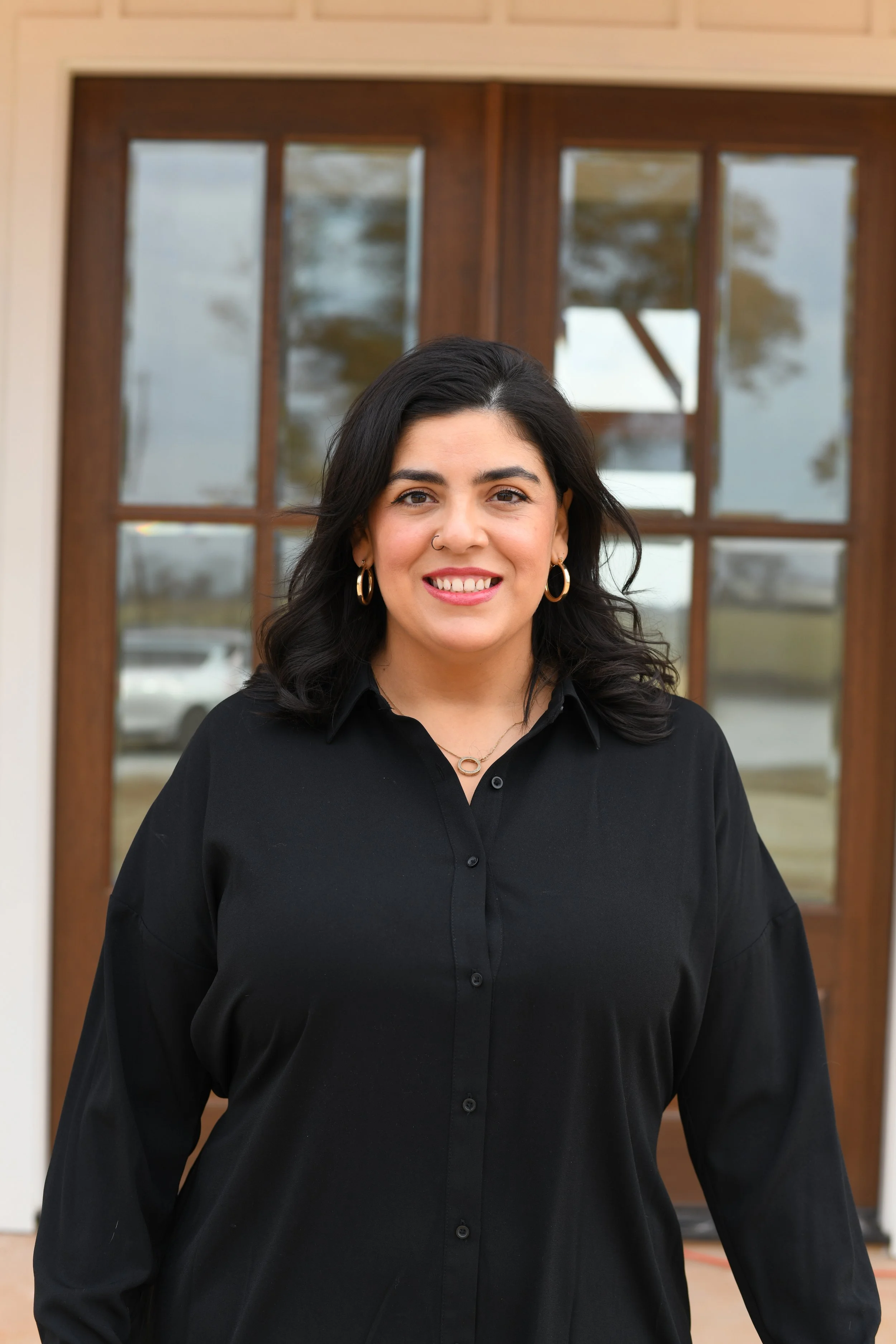 A woman with dark hair, wearing gold hoop earrings, a nose ring, and a black button-up shirt, standing in front of a wooden double door with glass panels. The background shows reflections of trees and a cloudy sky.
