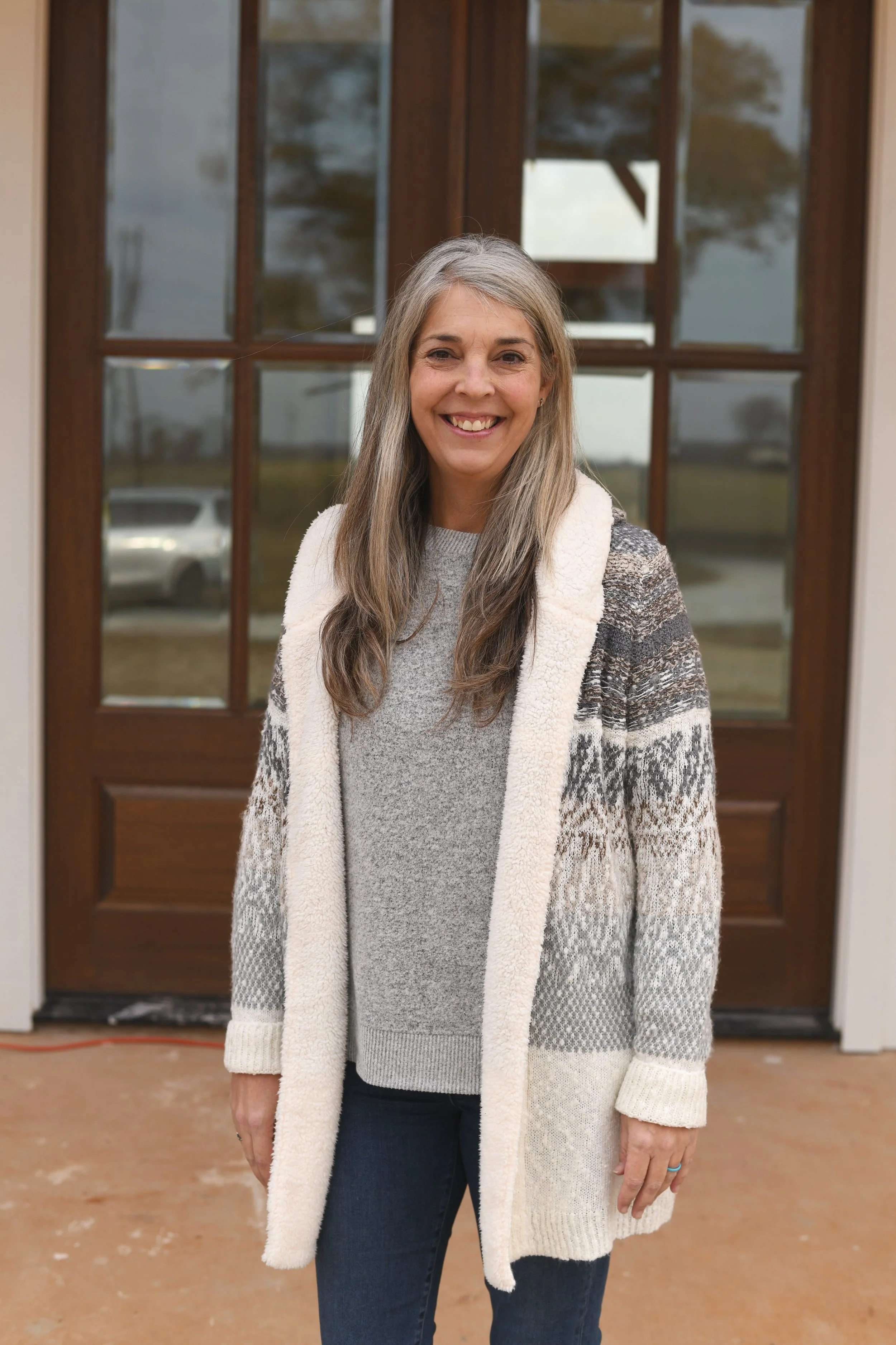 A woman with gray hair smiling at the camera, standing in front of a wooden door with glass panels, outdoors on a cloudy day.