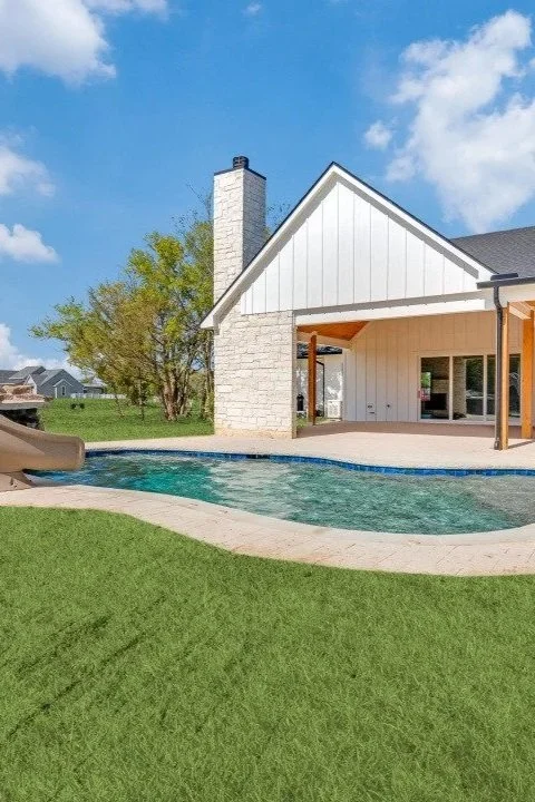 Backyard with swimming pool, slide, and patio area next to a house with white siding and a brick chimney, set against a bright blue sky with some clouds.