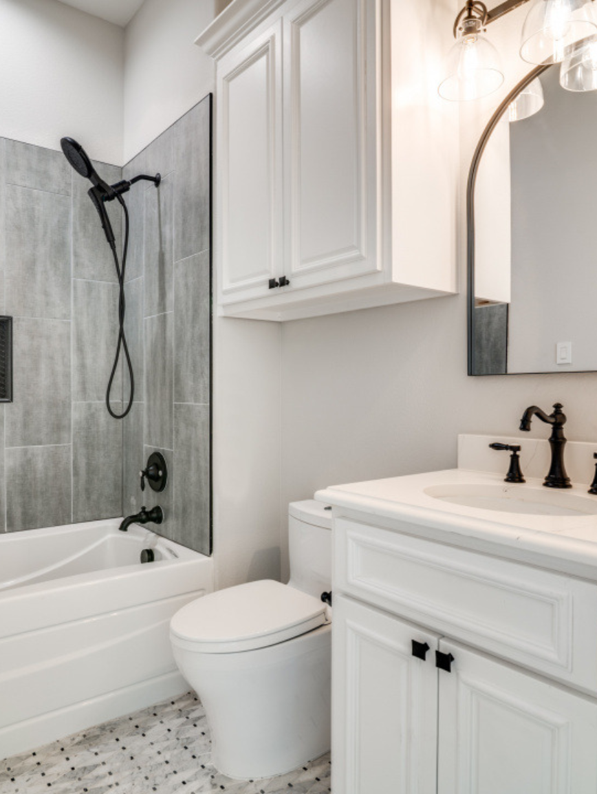 A bathroom with a shower/tub combination, white toilet, white vanity with black fixtures, wall-mounted cabinet, and a mirror with pendant lighting.