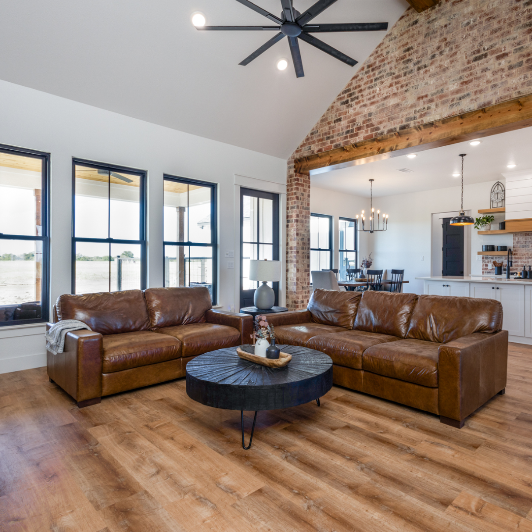 Living room with leather couch, coffee table, and large windows, open to a dining area and kitchen with exposed brick wall.
