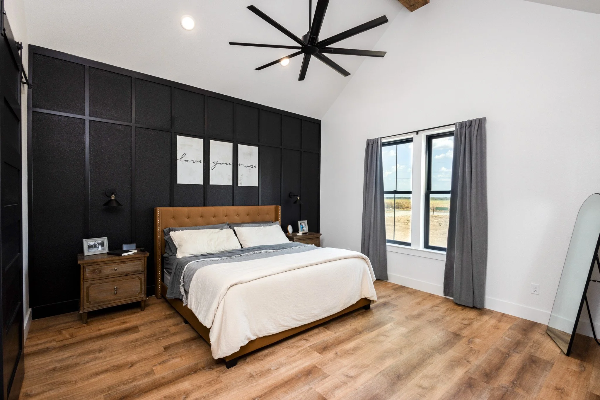 A modern bedroom with a black accent wall, a bed with tan headboard, white and gray bedding, and two wooden nightstands with framed photos. There's a ceiling fan, two windows with gray curtains, and a full-length mirror leaning against the wall.