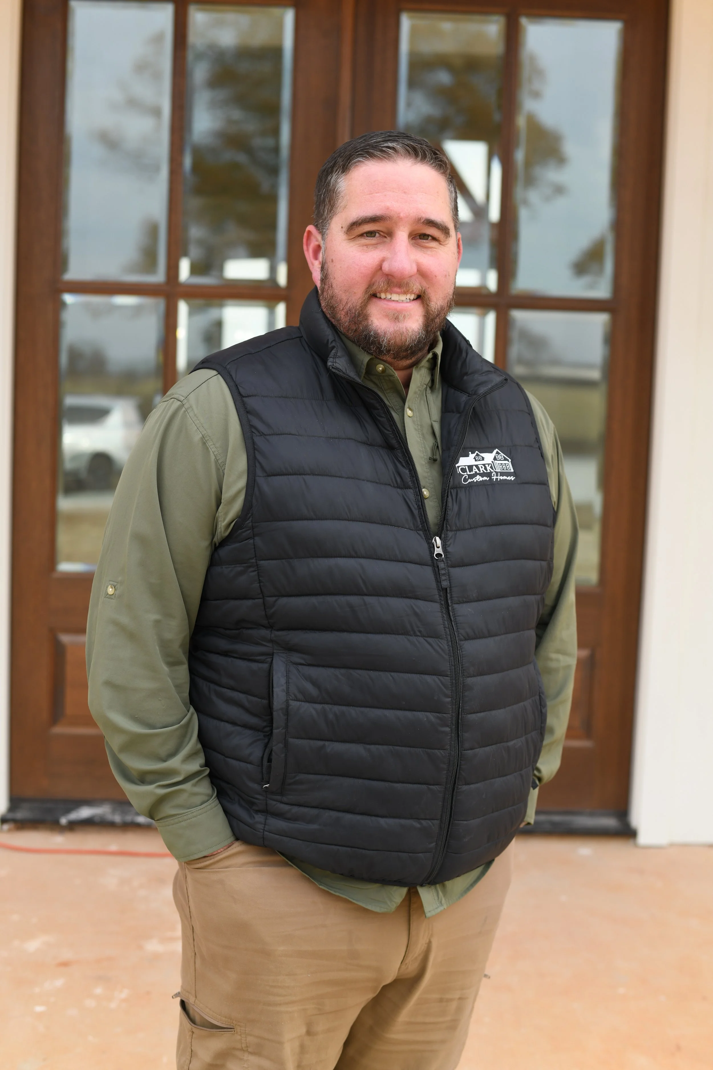 A man with a beard and short hair smiling, wearing a green shirt and a black quilted vest with a logo that says 'Clark Hubble Custom Homes', standing in front of a wooden door with glass panels.