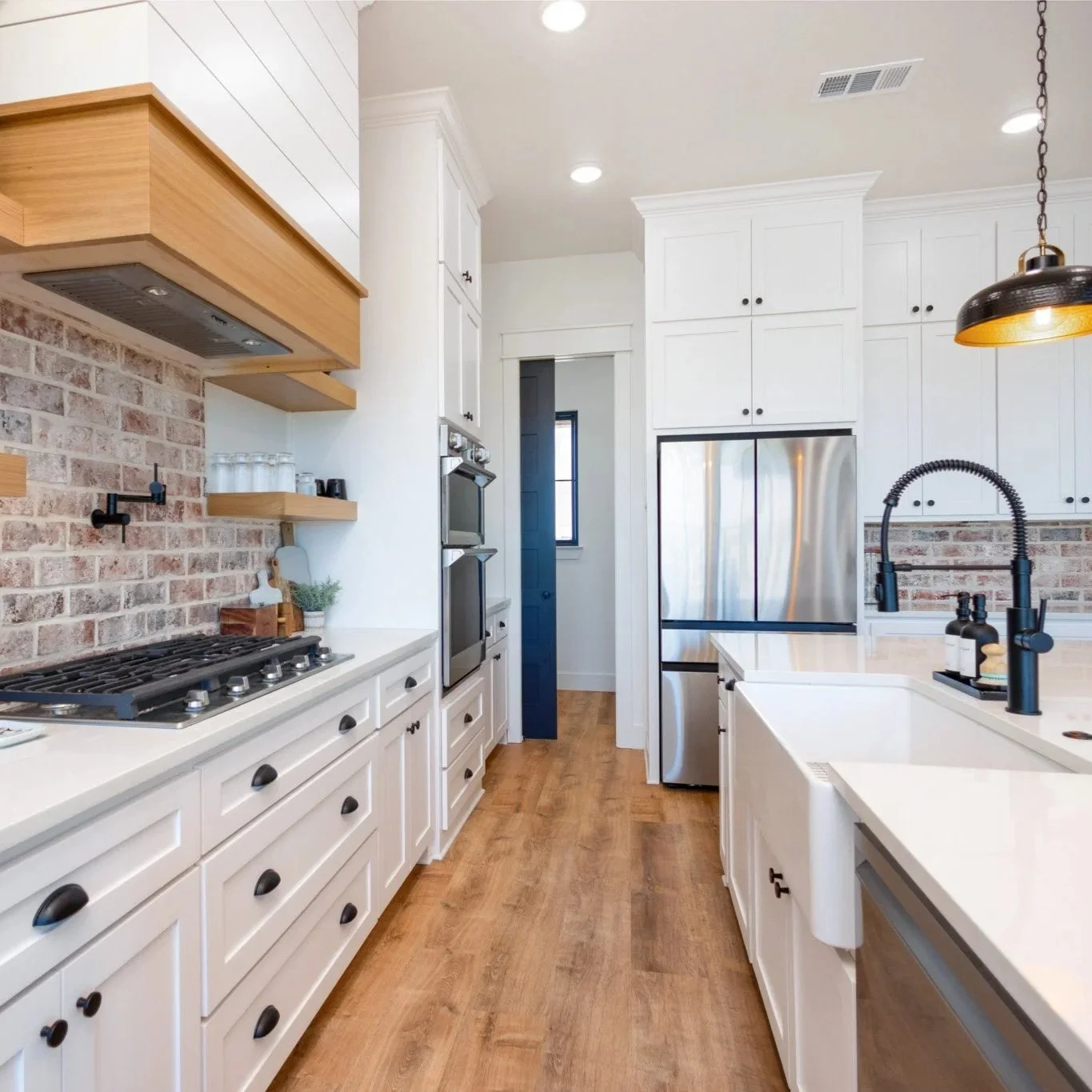 A modern kitchen with white cabinets, a brick backsplash, wooden shelves, stainless steel appliances, a black faucet, and a white farm sink. There's a black dome pendant light and wooden flooring.