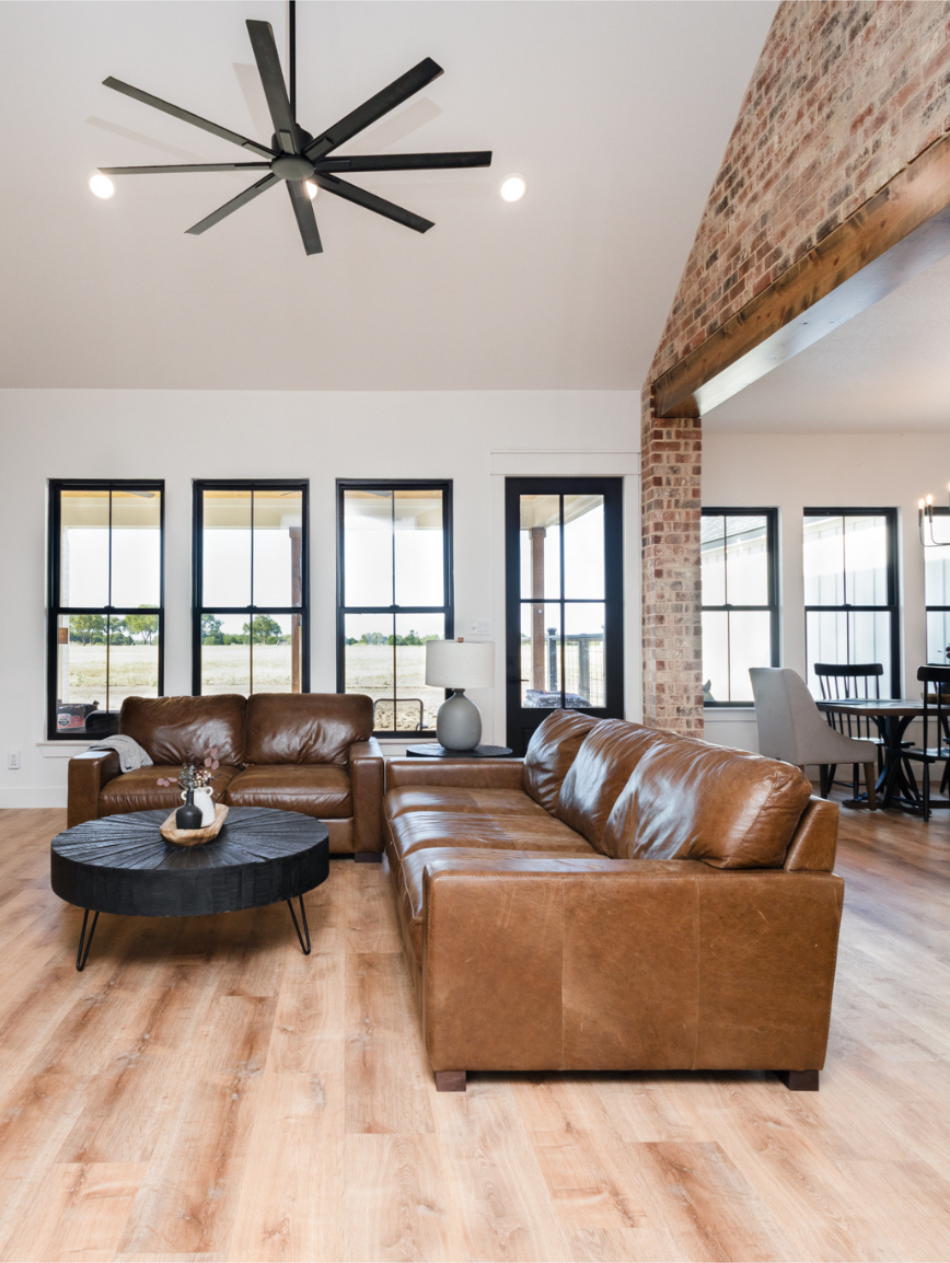Living room with brown leather sofas, black round coffee table, white lamp, large windows, brick wall, wooden flooring, and ceiling fan.