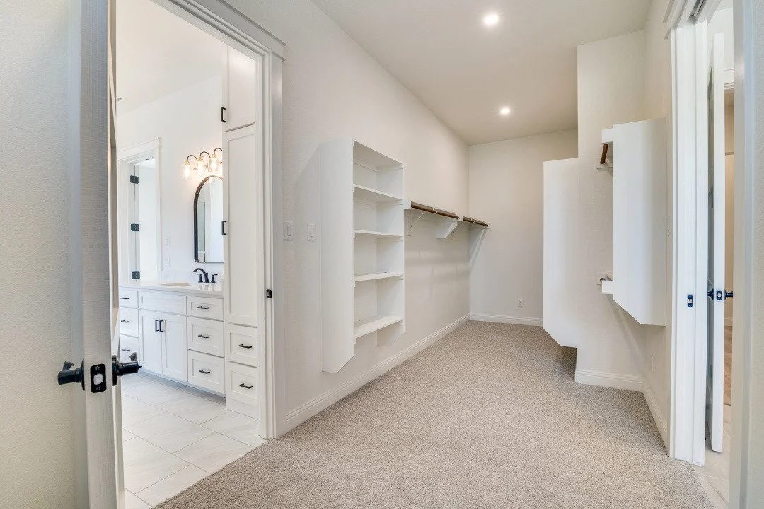 Empty walk-in closet with built-in shelves and hanging rods, adjacent to a bathroom with white cabinets and a mirror.