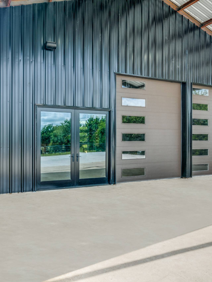 Exterior of a metal industrial building with black corrugated metal siding, glass double doors, and a garage door with multiple horizontal windows.