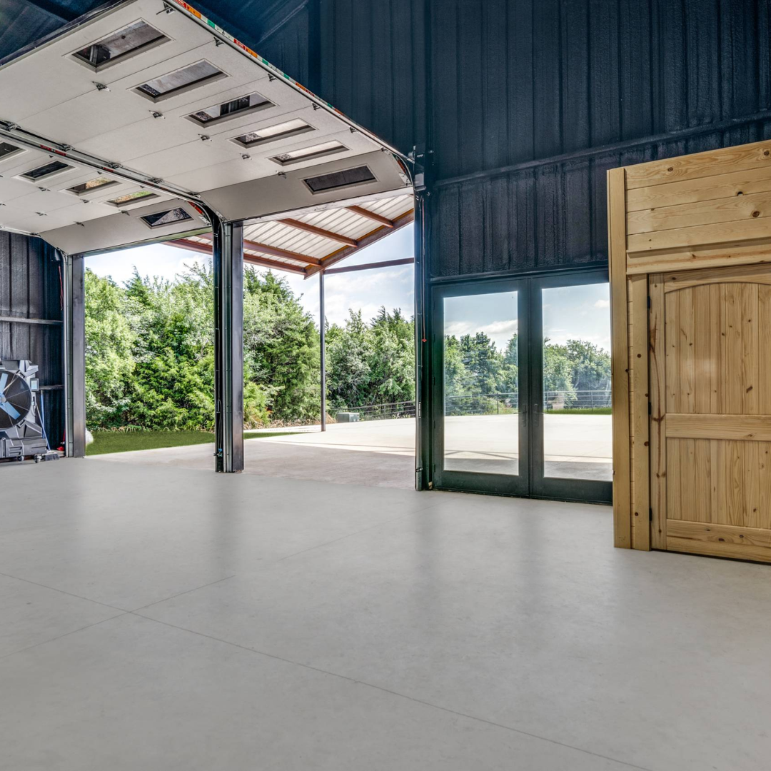 Empty garage with open doors, concrete floor, dark exterior walls, and a view of trees outside.