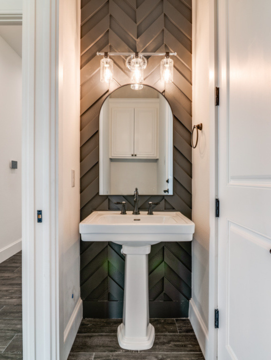 Bathroom with a white pedestal sink, arched mirror, and modern light fixture on dark wood-paneled wall.