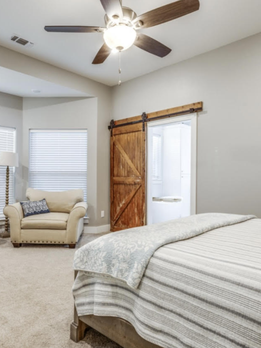A bedroom with a ceiling fan, a beige armchair with a patterned pillow, a window with blinds, and a wooden sliding door leading to a balcony or patio.